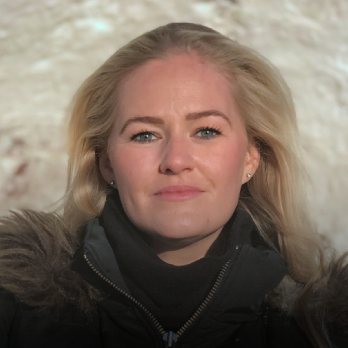 A close-up portrait of a woman with blonde hair and blue eyes, wearing a black jacket with a fur-lined hood, against a light-colored background.