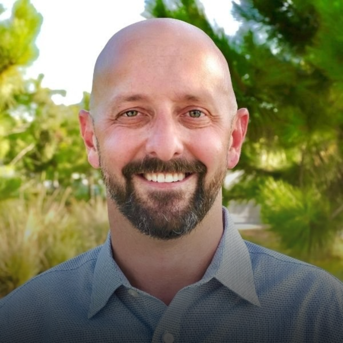 A smiling bald man with a beard and mustache, wearing a light blue checkered shirt, standing outdoors with green trees and foliage in the background.