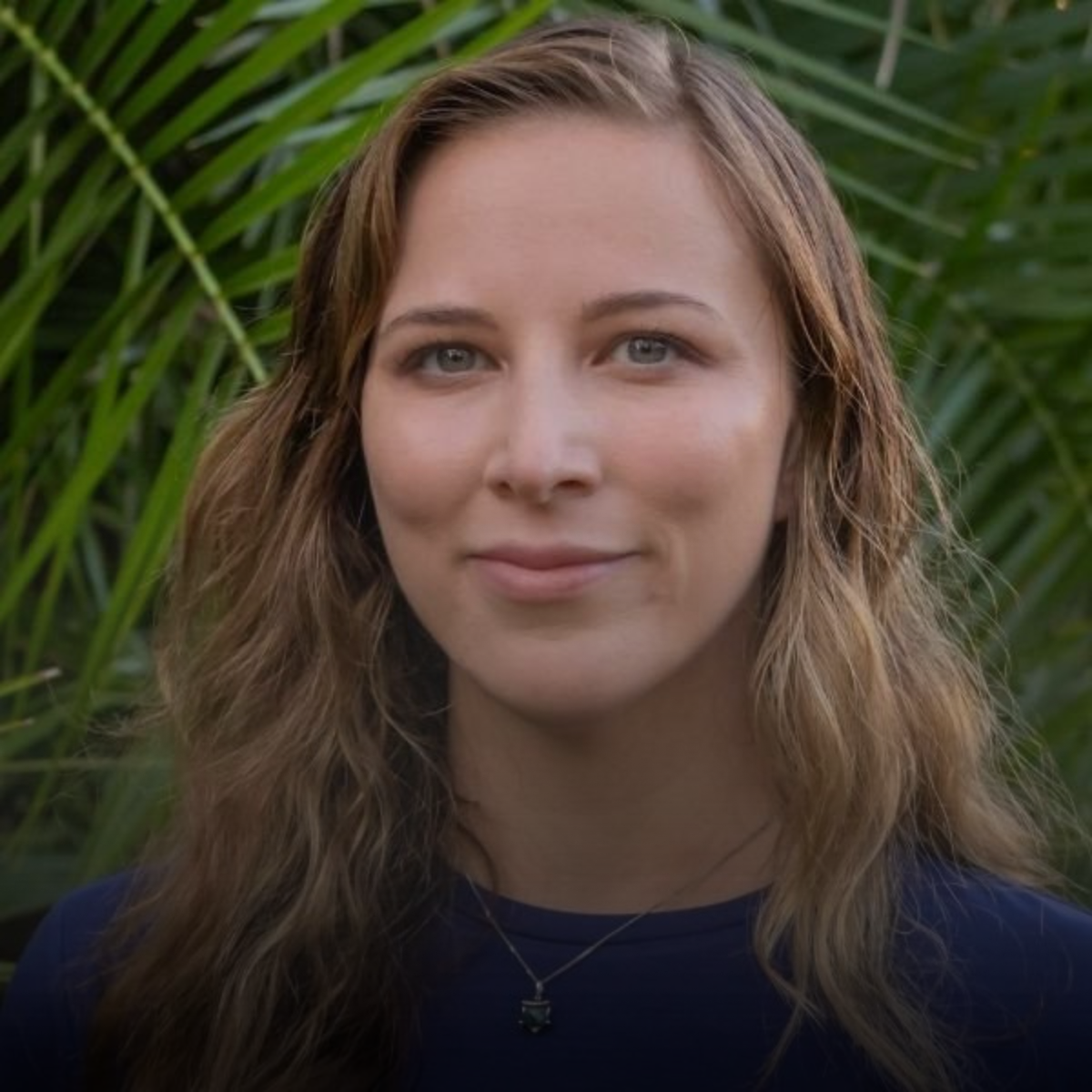 Close-up of a young woman with wavy, light brown hair and blue eyes, smiling slightly, with green palm leaves in the background.