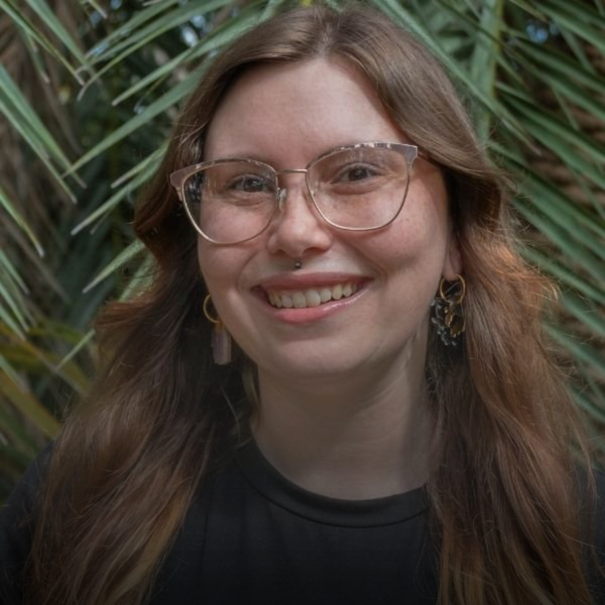 A young woman with light skin, glasses, and earrings smiling in front of green foliage.