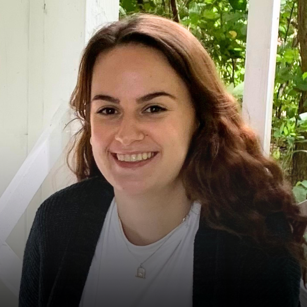 A woman with shoulder-length brown hair smiling outdoors near green foliage, wearing a white top, a black sweater, and a silver necklace.