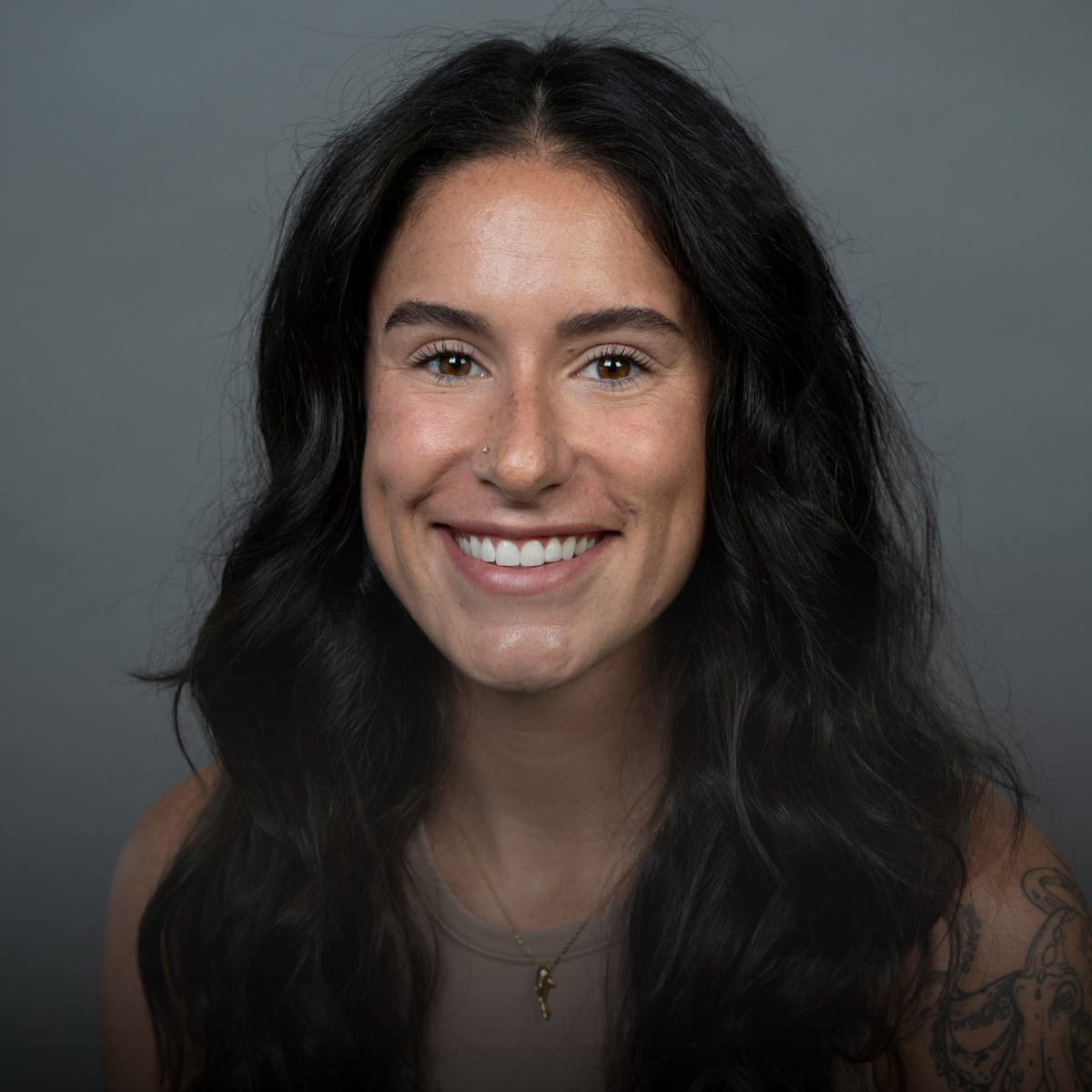 A woman with long, wavy dark hair, smiling, wearing a beige top and gold necklace, against a gray background.