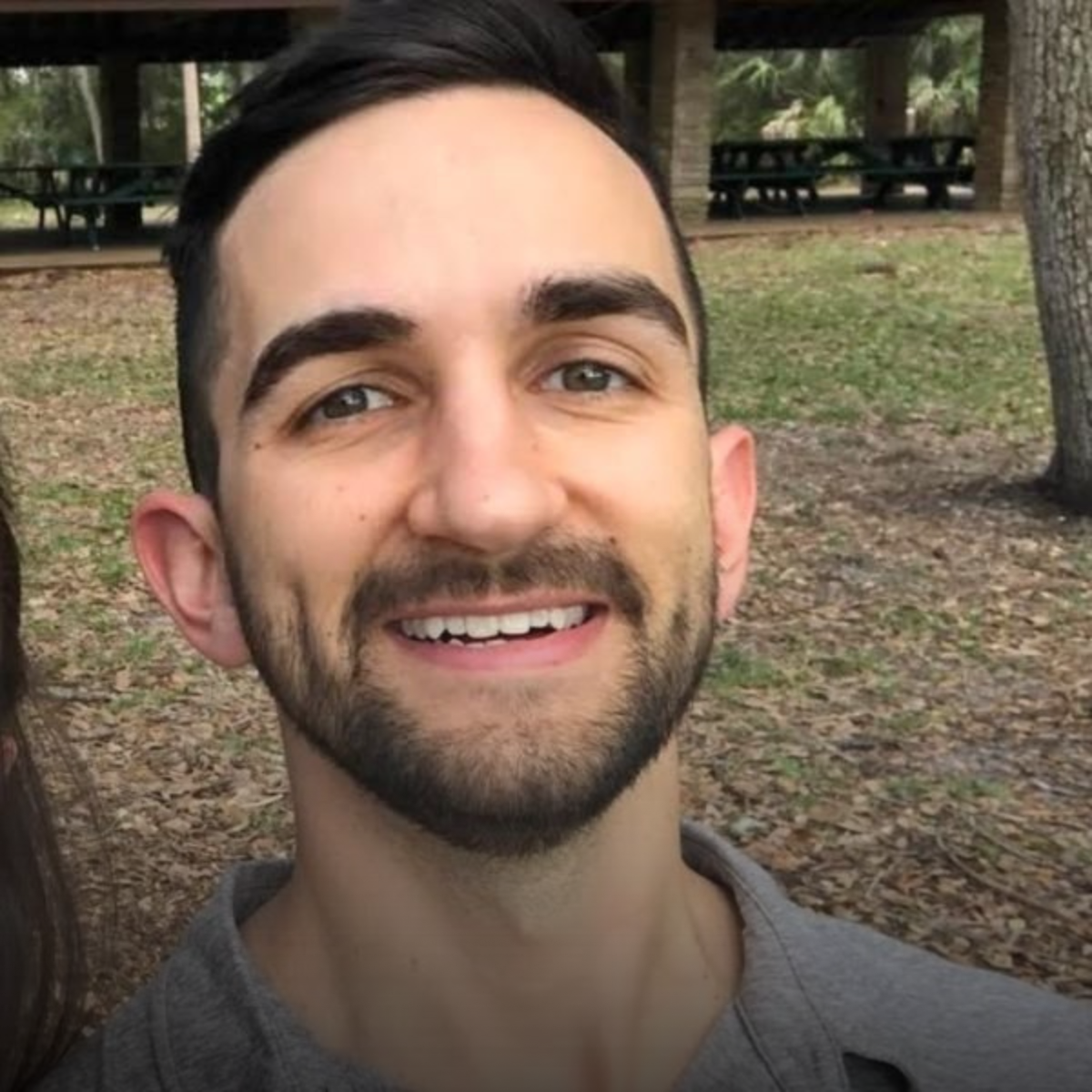 Close-up of a smiling young man with dark hair and a beard, outdoors in a park with trees and picnic tables in the background.