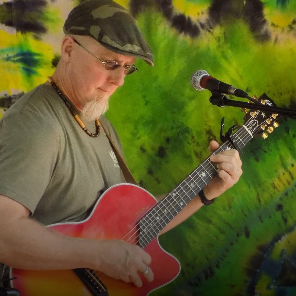 A man wearing a camouflage cap, sunglasses, and a green t-shirt playing a red acoustic guitar in front of a colorful, tie-dye style background.