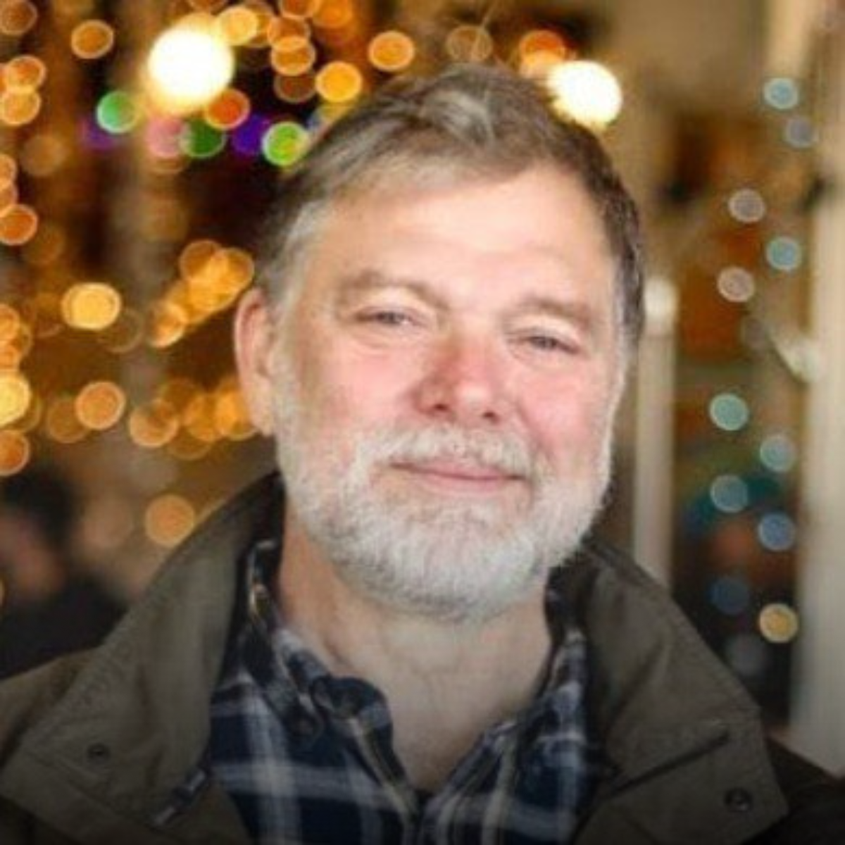 A middle-aged man with gray hair and a beard, smiling, with a background of blurred colorful Christmas lights.