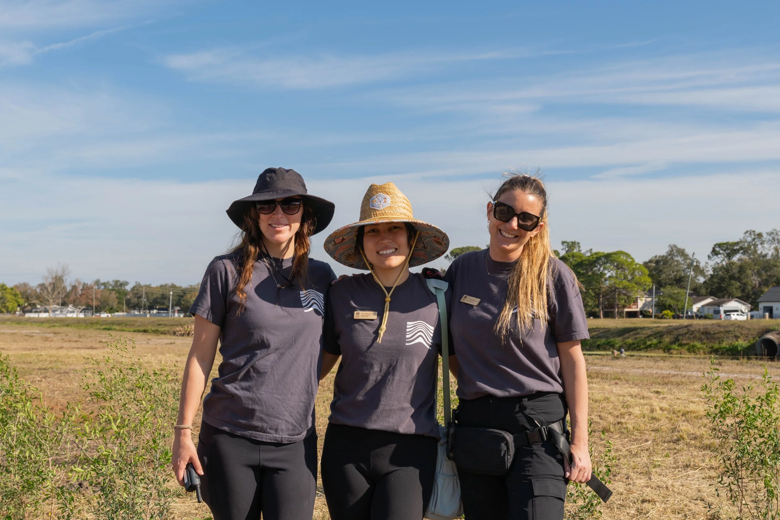 Three women standing outdoors on a sunny day, wearing hats and sunglasses, smiling at the camera, with trees, houses, and a blue sky with some clouds in the background.