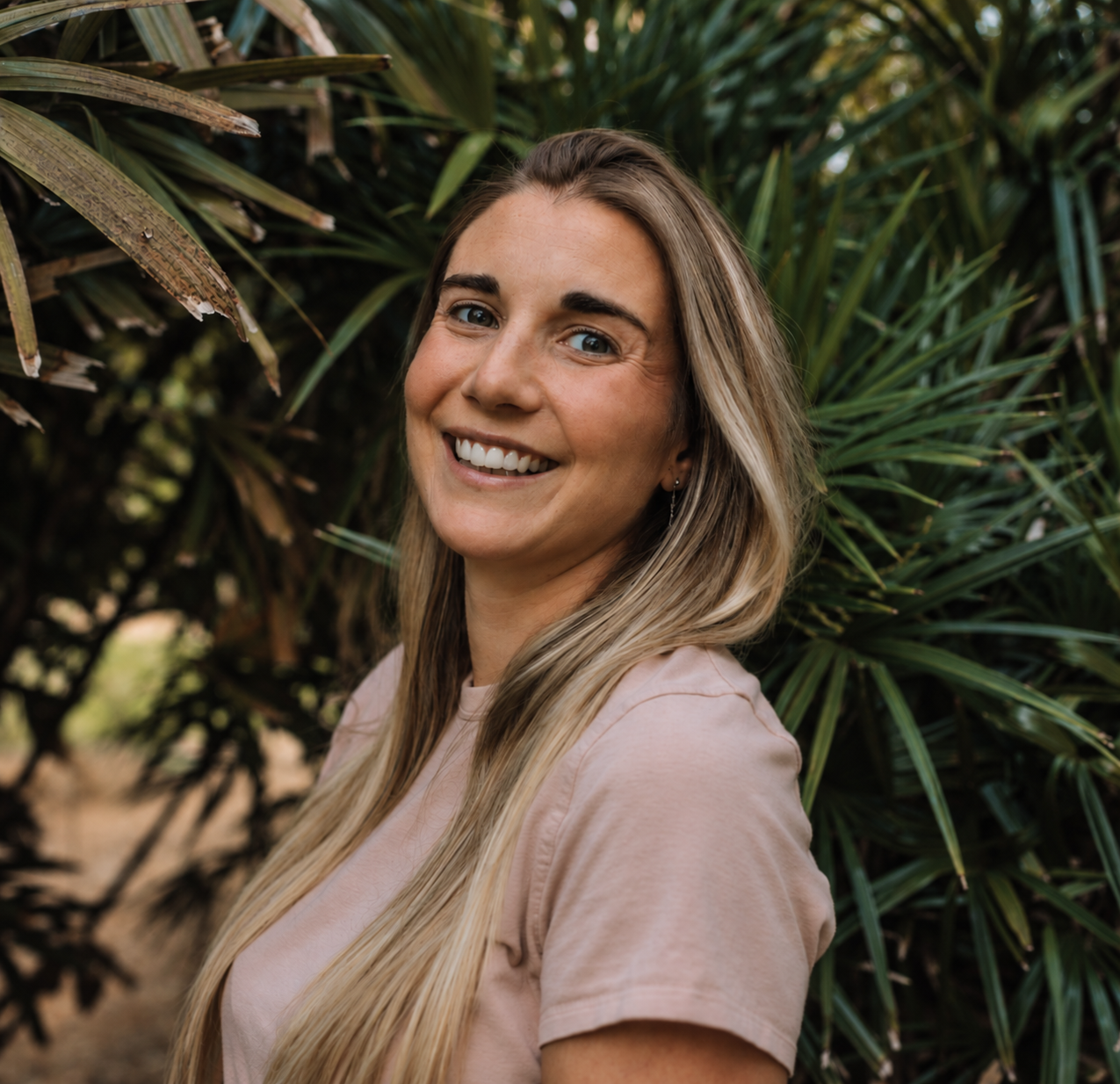 A woman with long blonde hair smiling in front of green leafy plants.