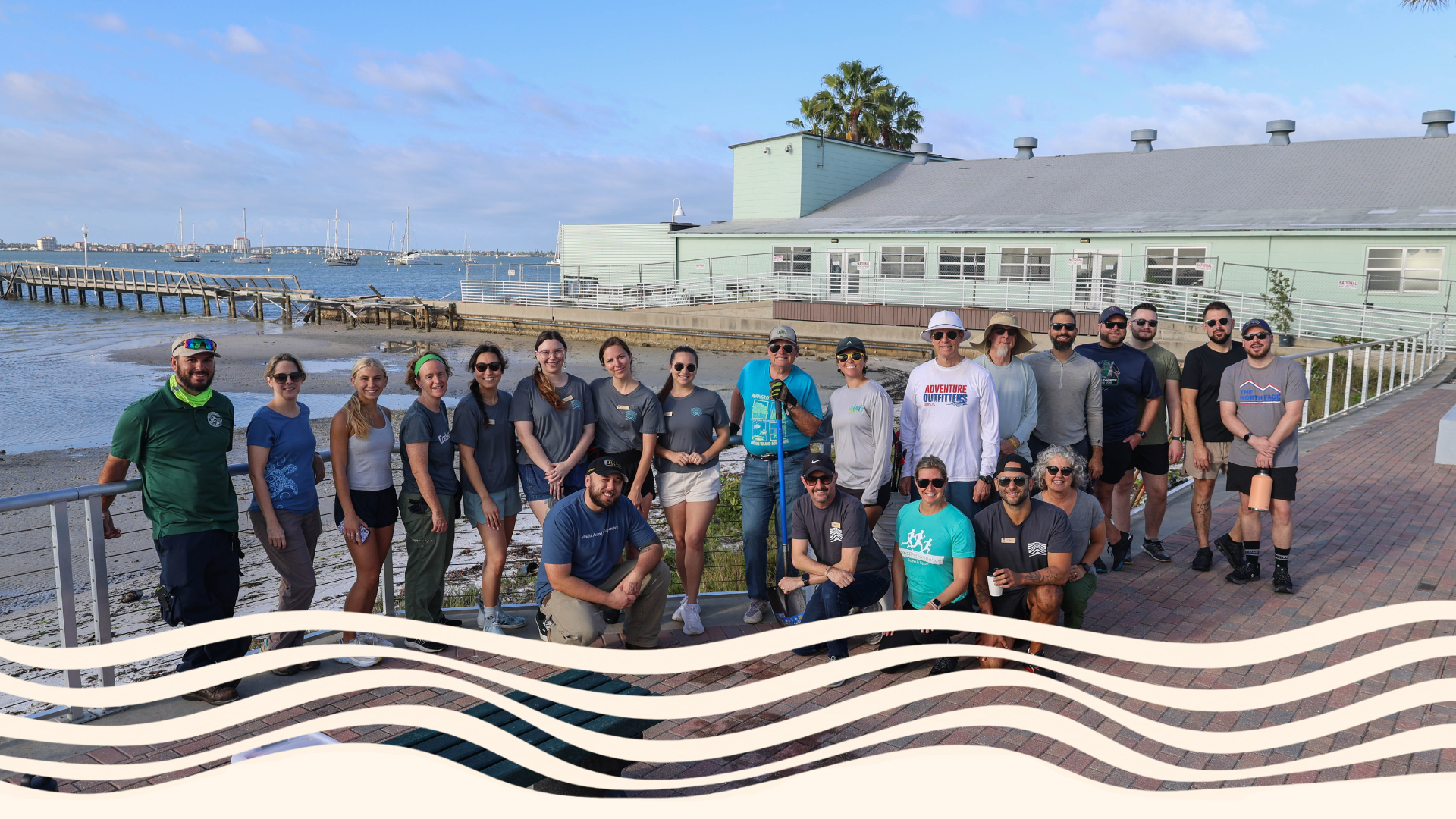 Group of people standing on a waterfront sidewalk, smiling for a photo with a marina and docked boats in the background on a partly cloudy day.