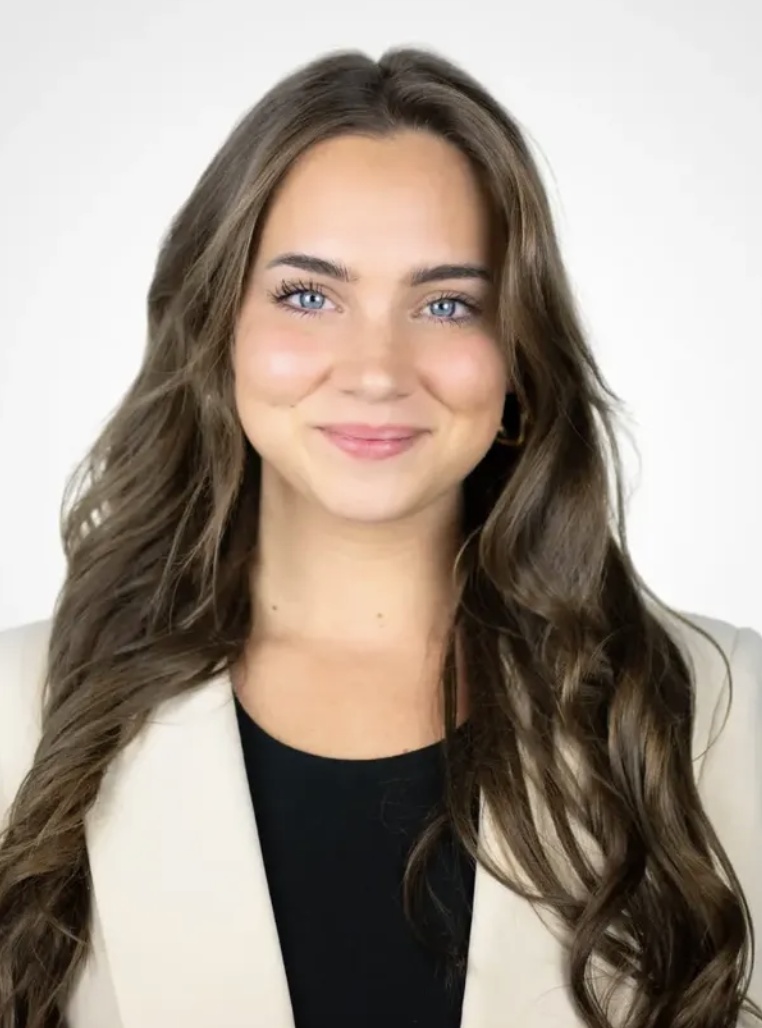Young woman with long wavy brown hair, blue eyes, and light skin, smiling, wearing a beige blazer over a black top, with a plain white background.