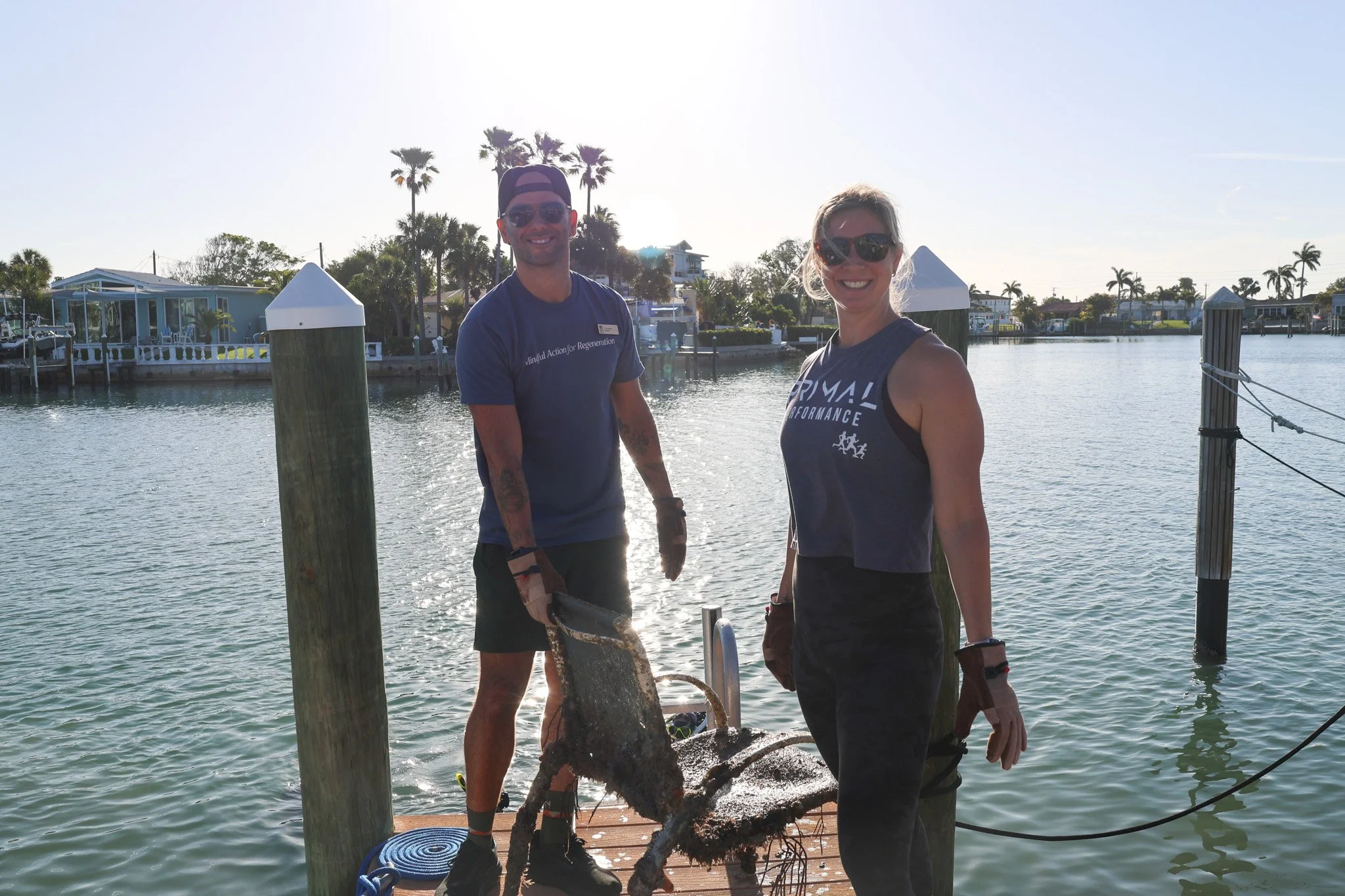 Two people on a dock by the water, with palm trees and houses in the background. One person is holding a large fish, and they are both smiling.