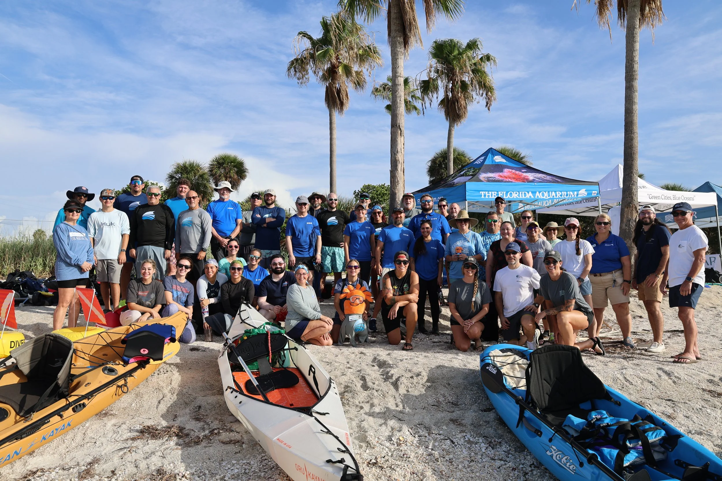 Group of people posing on a beach with kayaks and outdoor gear, under a blue sky with palm trees and a tent in the background.