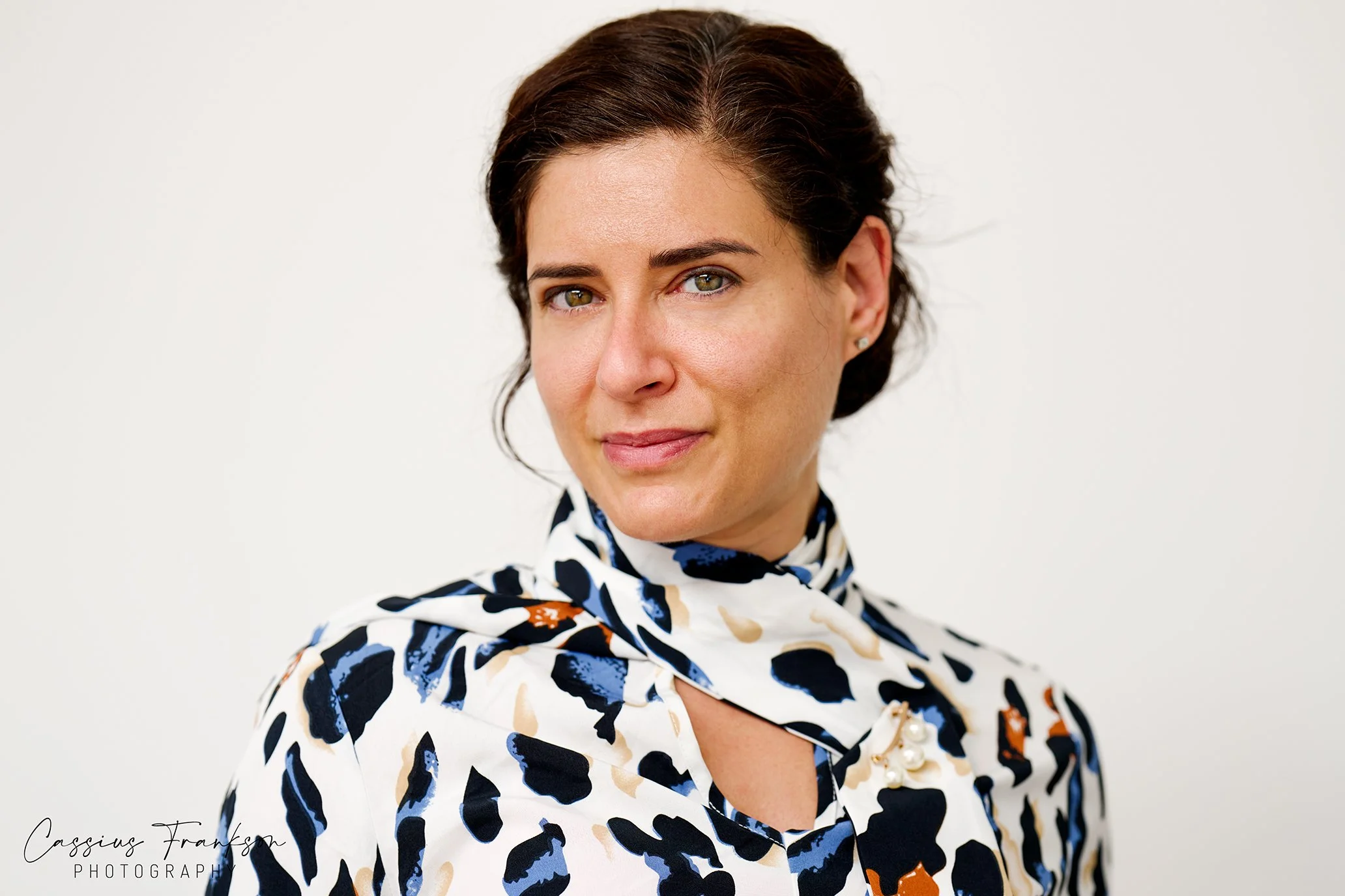 A woman with short brown hair and hazel eyes wearing a colorful patterned scarf or blouse with a floral design, posing against a plain white background.