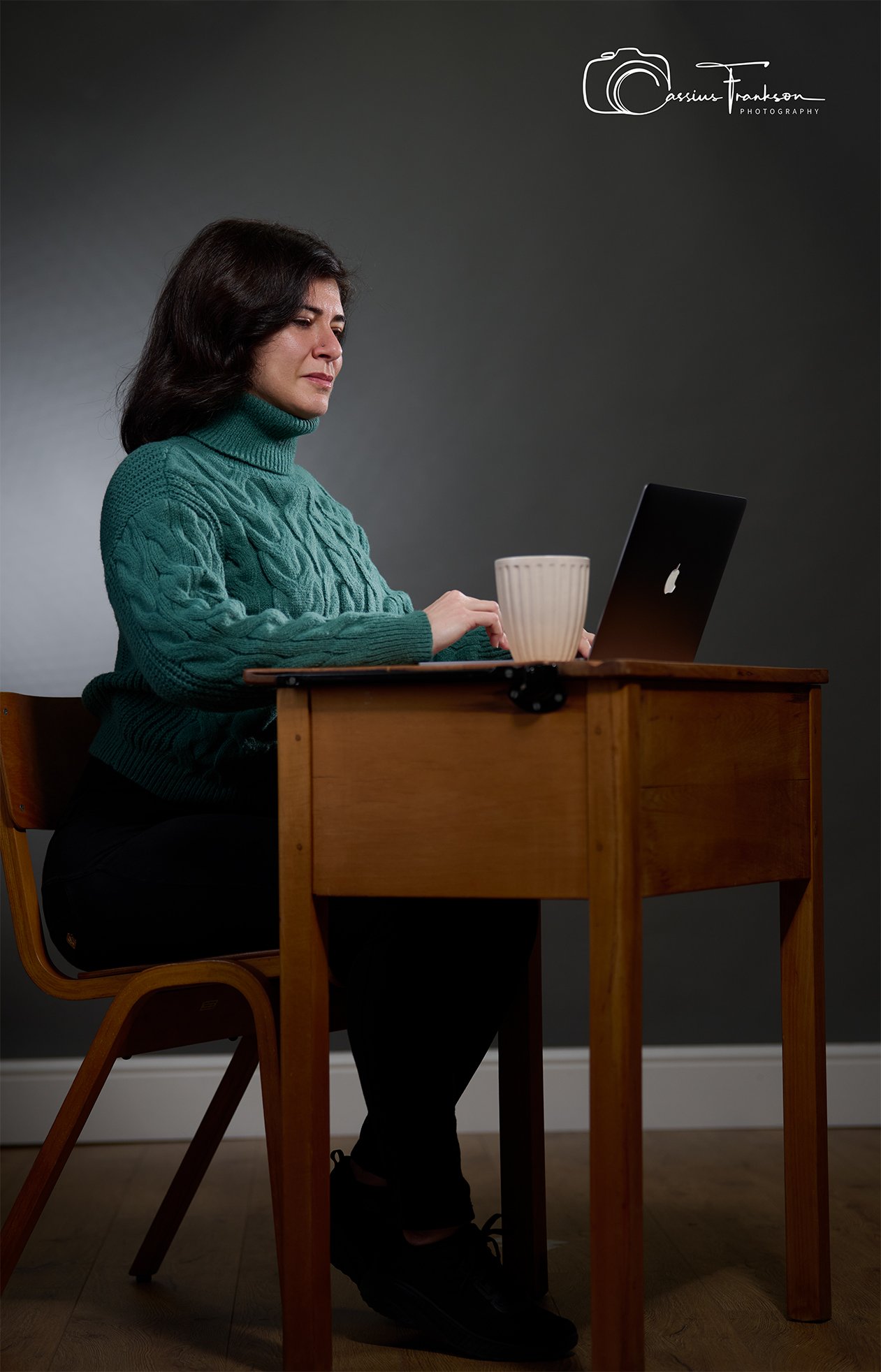 Woman with dark hair, wearing a teal cable-knit turtleneck sweater, sitting at a wooden table using a black laptop, with a white ceramic mug nearby, against a plain dark background.