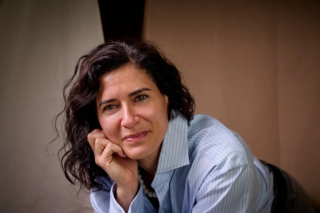 A woman with shoulder-length dark hair, wearing a blue striped shirt, leaning on one hand and looking at the camera with a subtle smile.