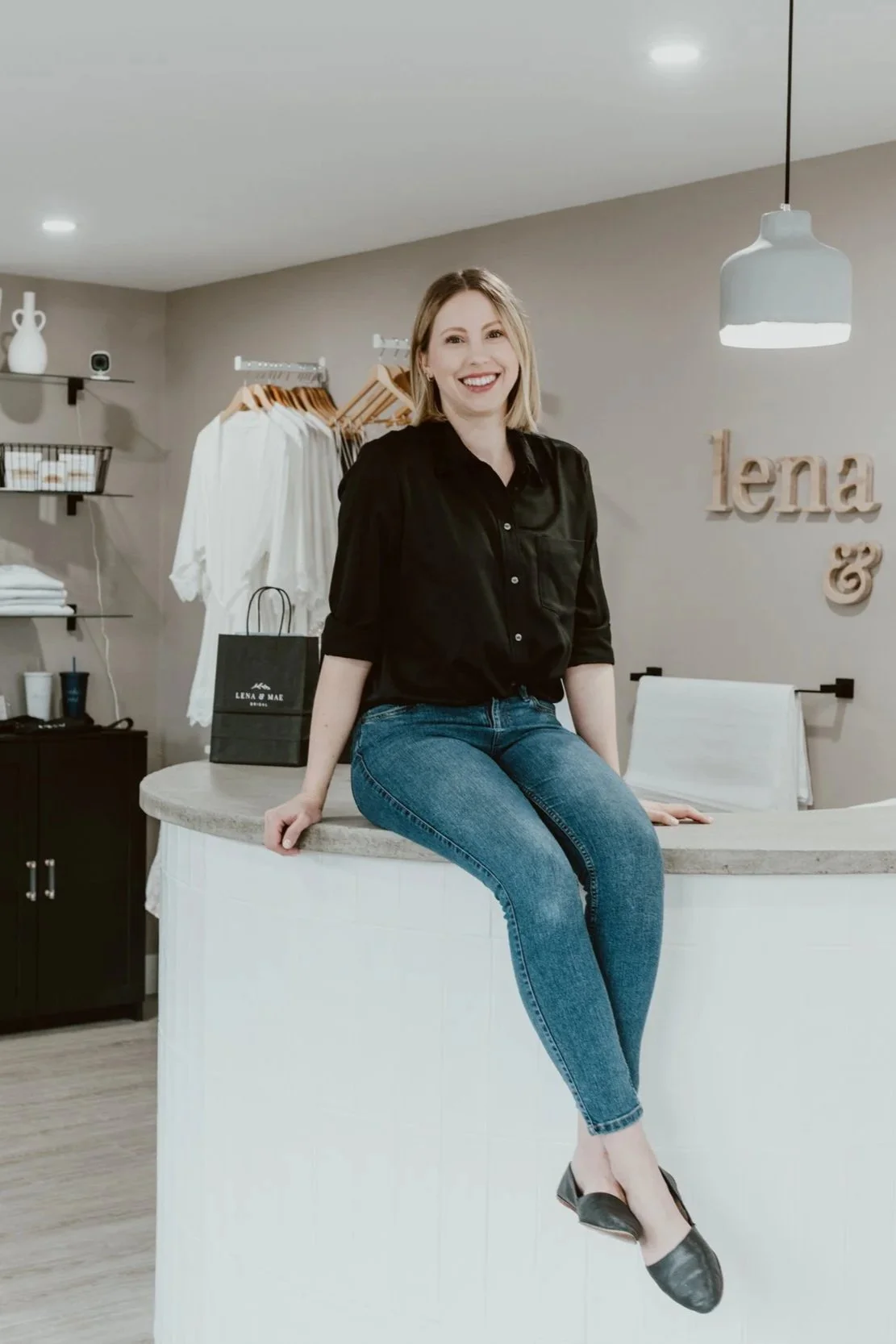A woman with blonde hair, wearing a black shirt and blue jeans, sitting on a curved counter inside a clothing store with various garments and accessories in the background.