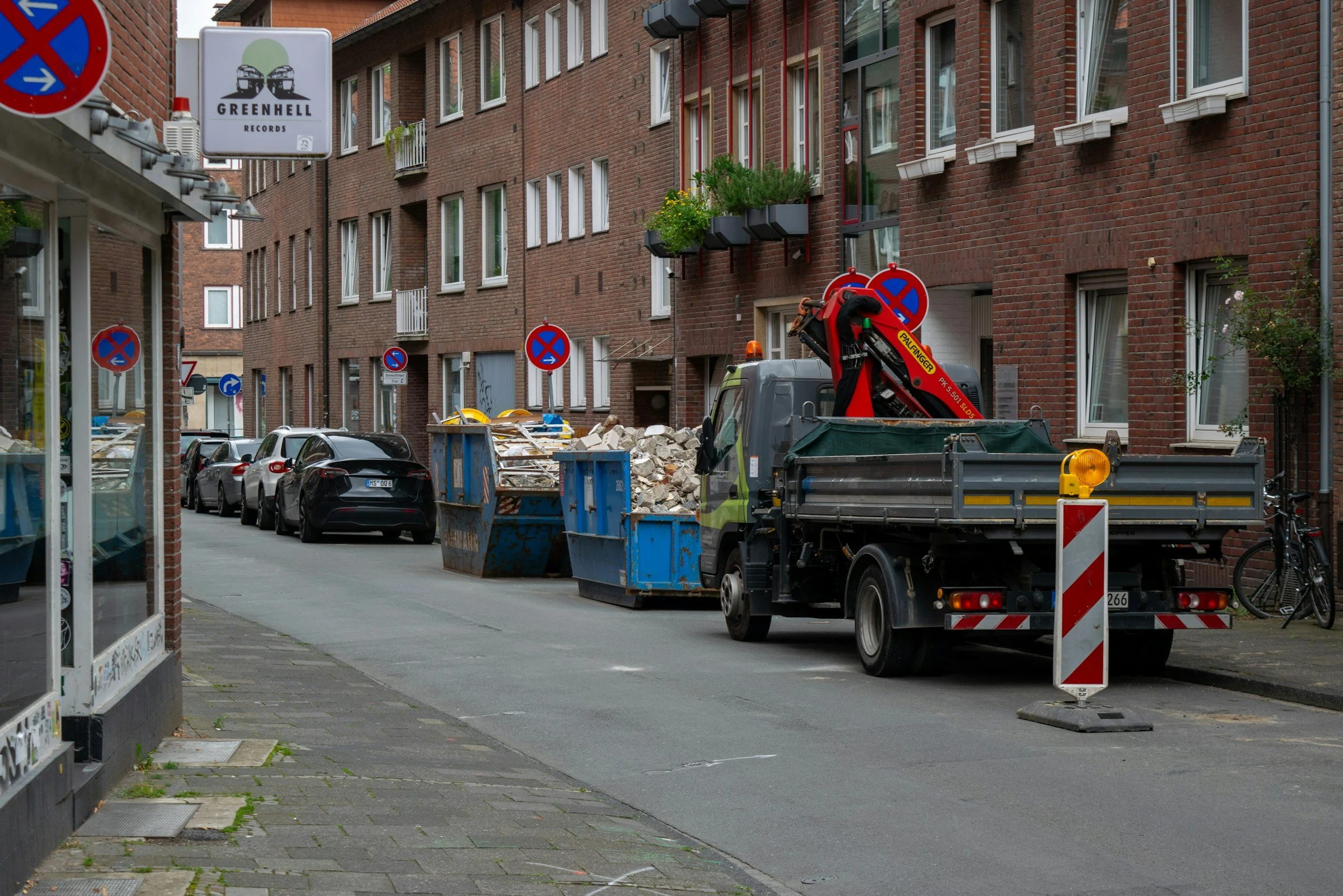 Straßenszene mit Bauarbeiten. Es ist eine mehrstöckige Wohnanlage aus roten Ziegelsteinen zu sehen. Ein Baukran und Müllcontainer sind auf der Straße, sowie parkende Autos. Verkehrsschilder verbieten Halten und Parken.