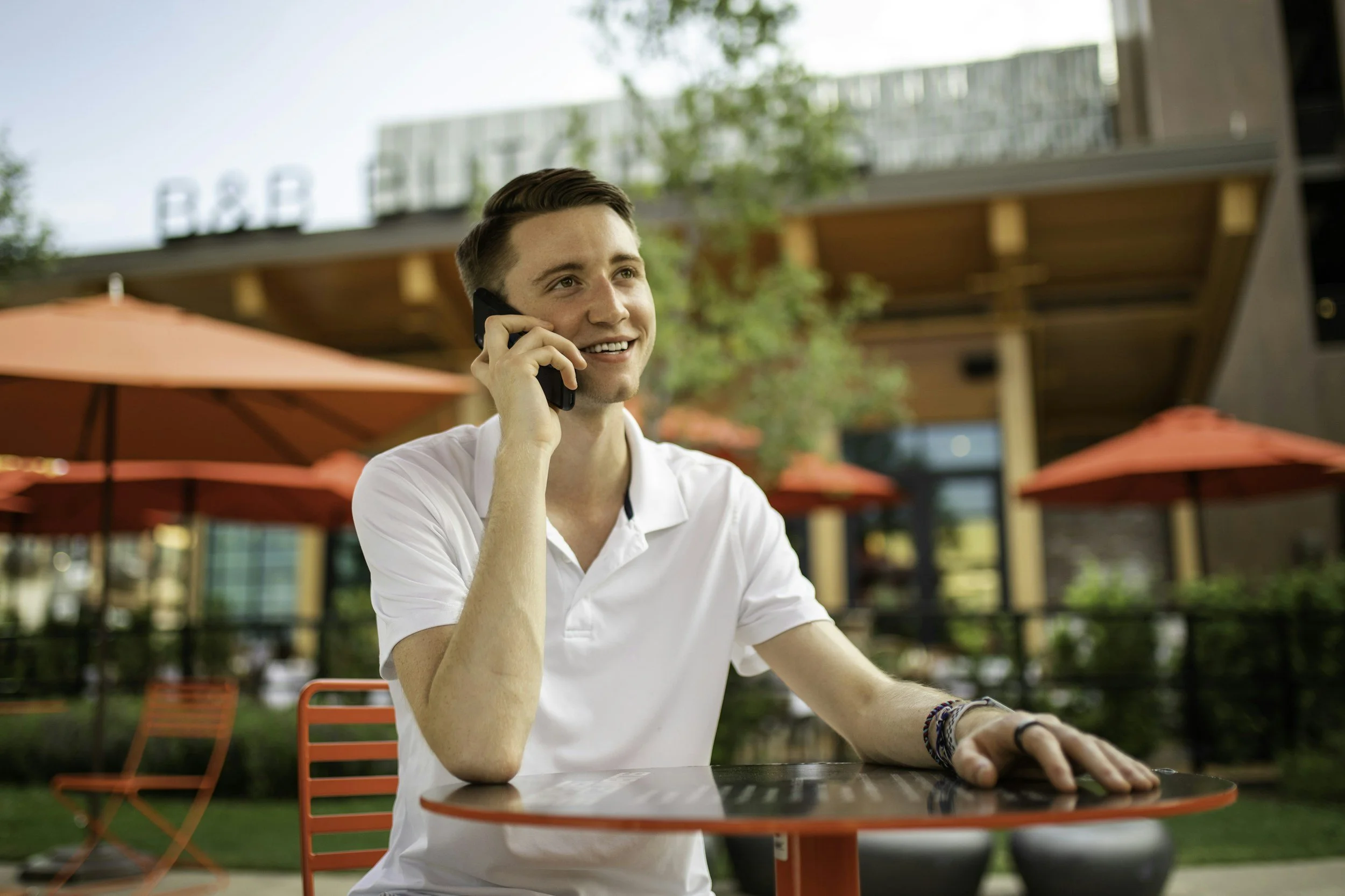 A young man with short hair, smiling, talking on a cellphone while sitting outdoors at a red table in a restaurant patio area with red umbrellas and a wooden building in the background.