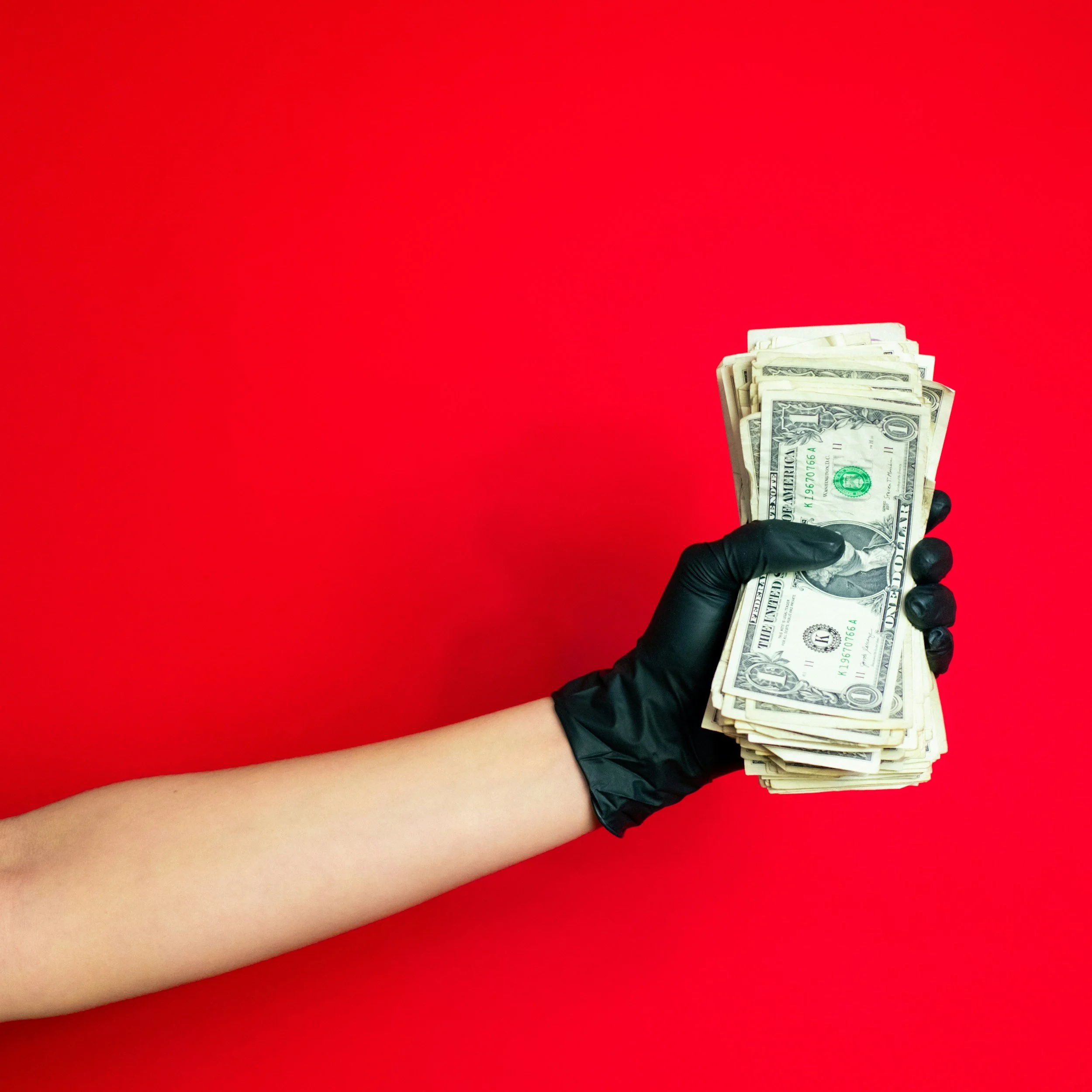 A hand wearing a black glove holding a bunch of U.S. dollar bills against a bright red background.