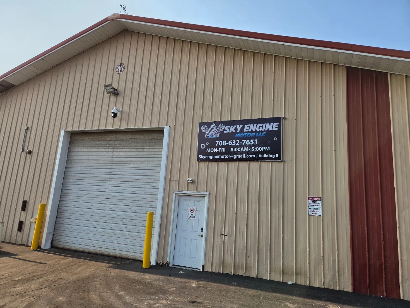Exterior view of a beige metal building with a large garage door, a small door, and signage for Sky Engine Motor LLC, including contact information and operating hours, with yellow safety posts on either side of the garage door.