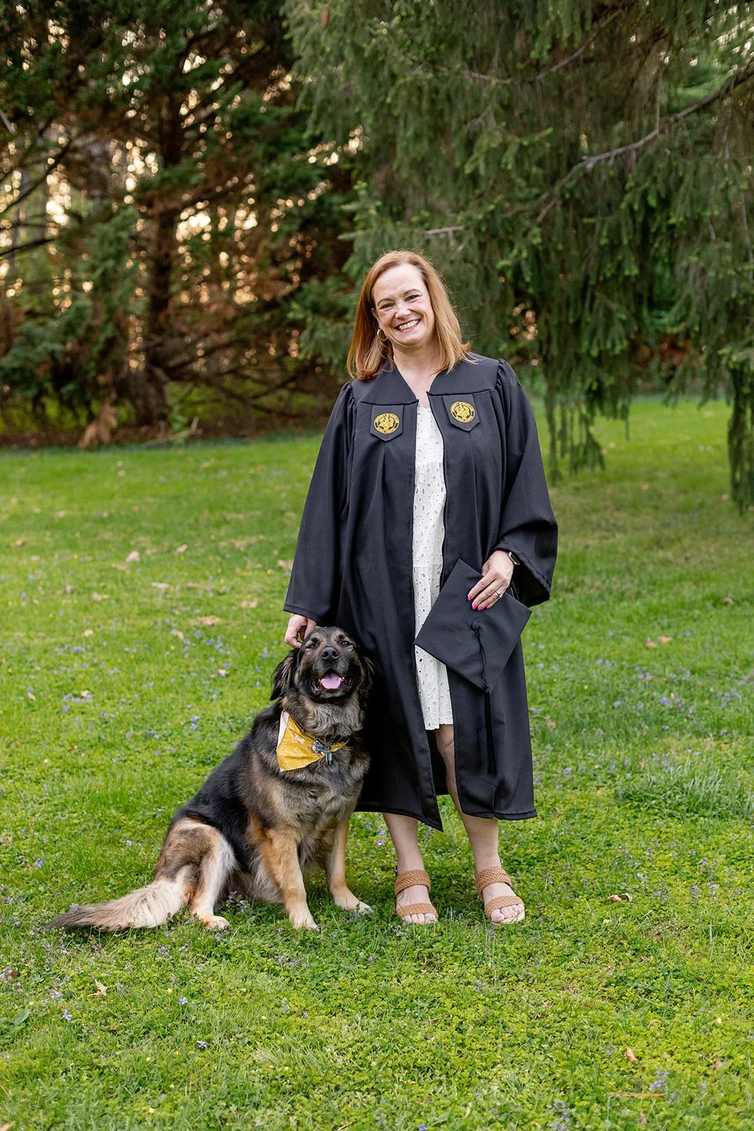 A woman in a graduation gown holding a cap stands outdoors next to a large dog wearing a bandana, with trees and grass in the background.