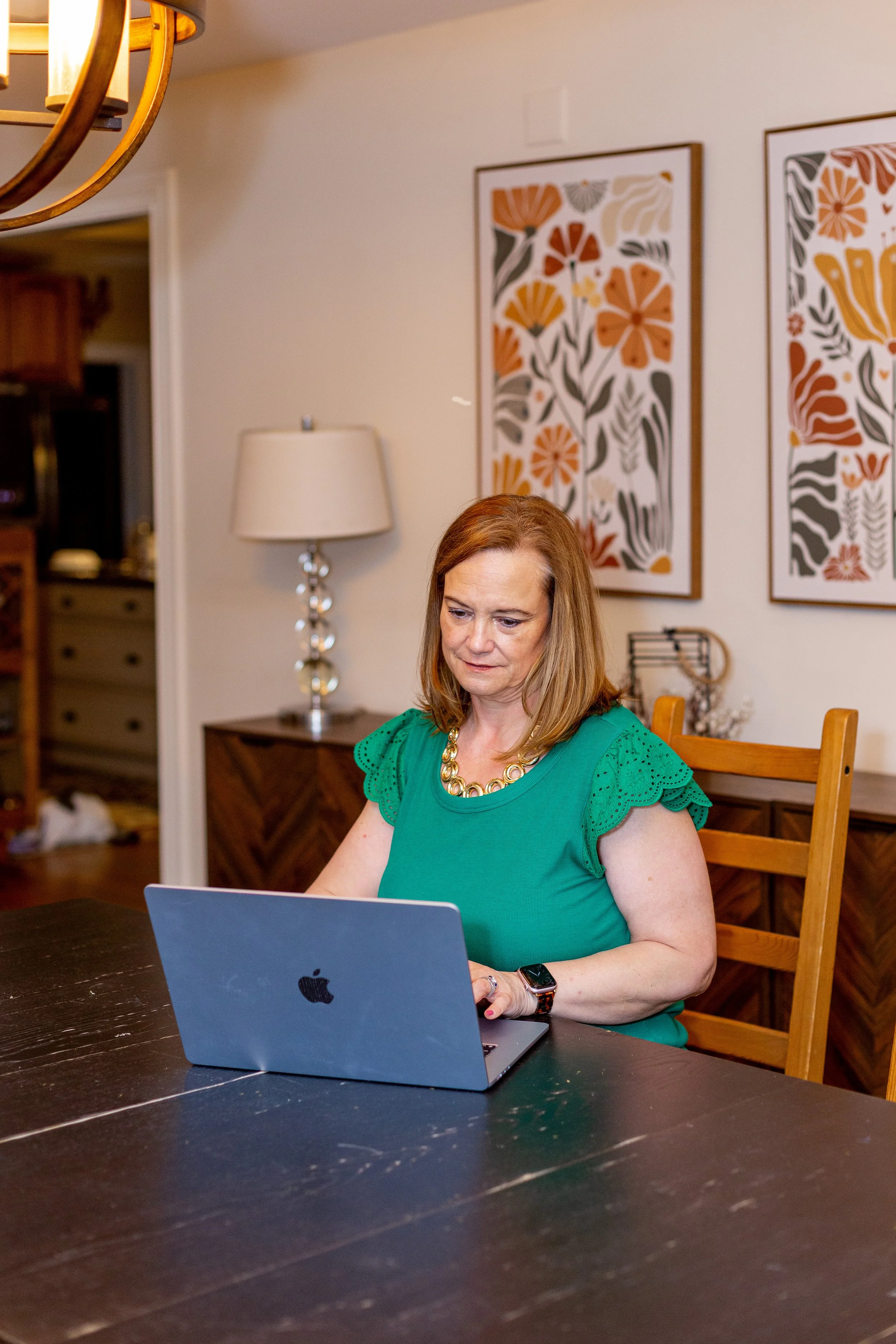 A woman with shoulder-length reddish-brown hair wearing a green top with eyelet details on the sleeves, sitting at a dark wood table and working on a silver MacBook. The background features framed floral artwork, a lamp with a white shade and metallic base, and a glimpse of a kitchen area.