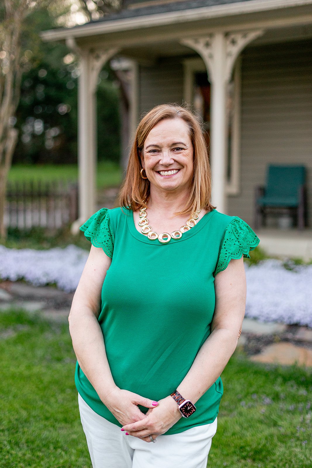 A woman with shoulder-length red hair wearing a green top with lace sleeves, a gold necklace, and a watch, smiling outdoors in front of a house and garden.