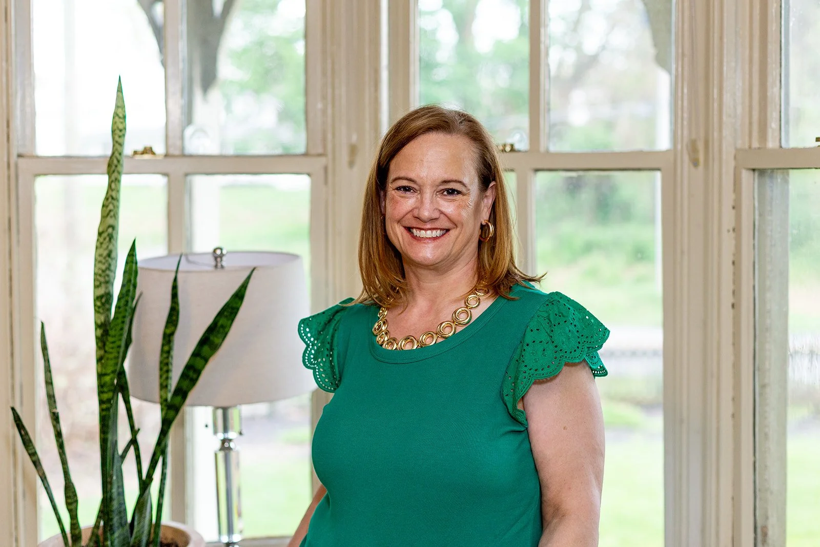A woman with shoulder-length red hair smiling, wearing a green top with decorative sleeves and a gold necklace, standing inside near windows and a potted plant.