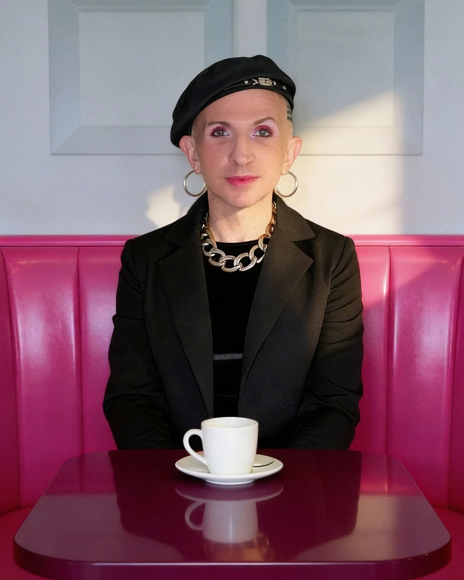 Slay Channel founder, Remy JF Thompson, wearing hoop earrings, a chunky chain necklace, a black beret, black blazer, sitting in a pink booth at a diner with a white coffee cup on the table