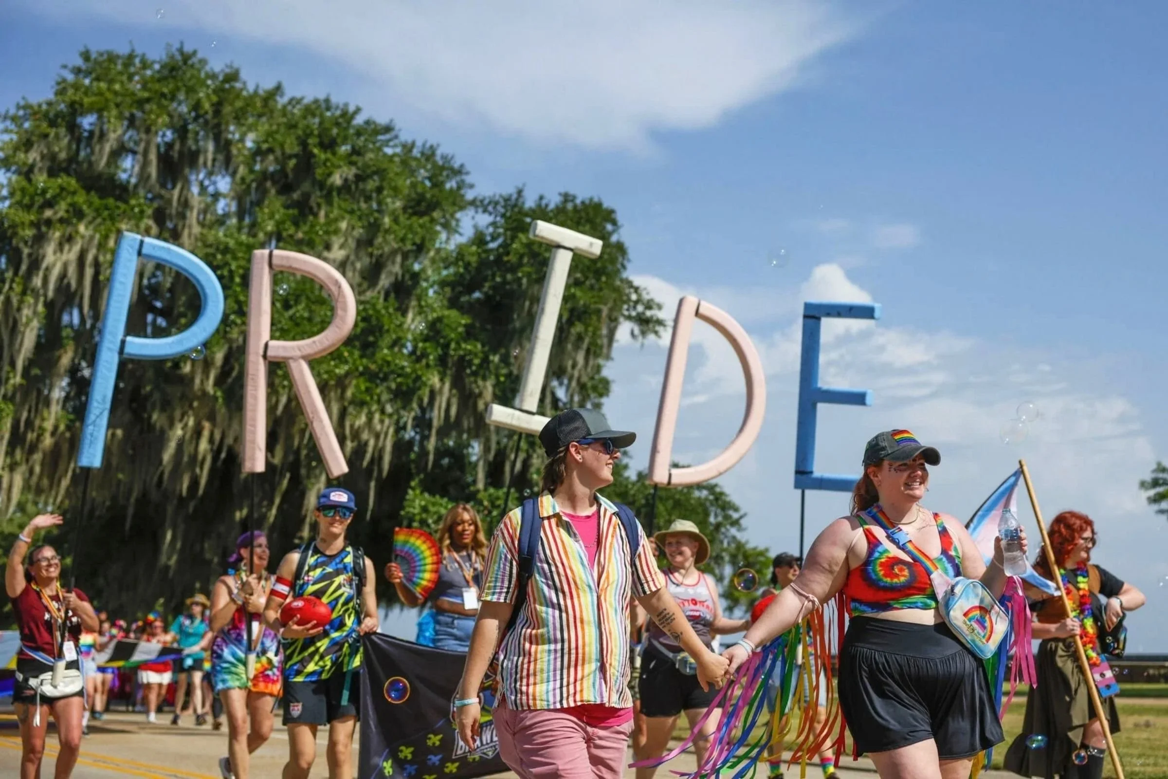 A group of people participating in a Pride parade, holding hands and wearing colorful rainbow clothing and accessories. There are large pastel-colored letters spelling out 'PRIDE' in the background, and some people are holding pride flags and wearing rainbow-themed items.