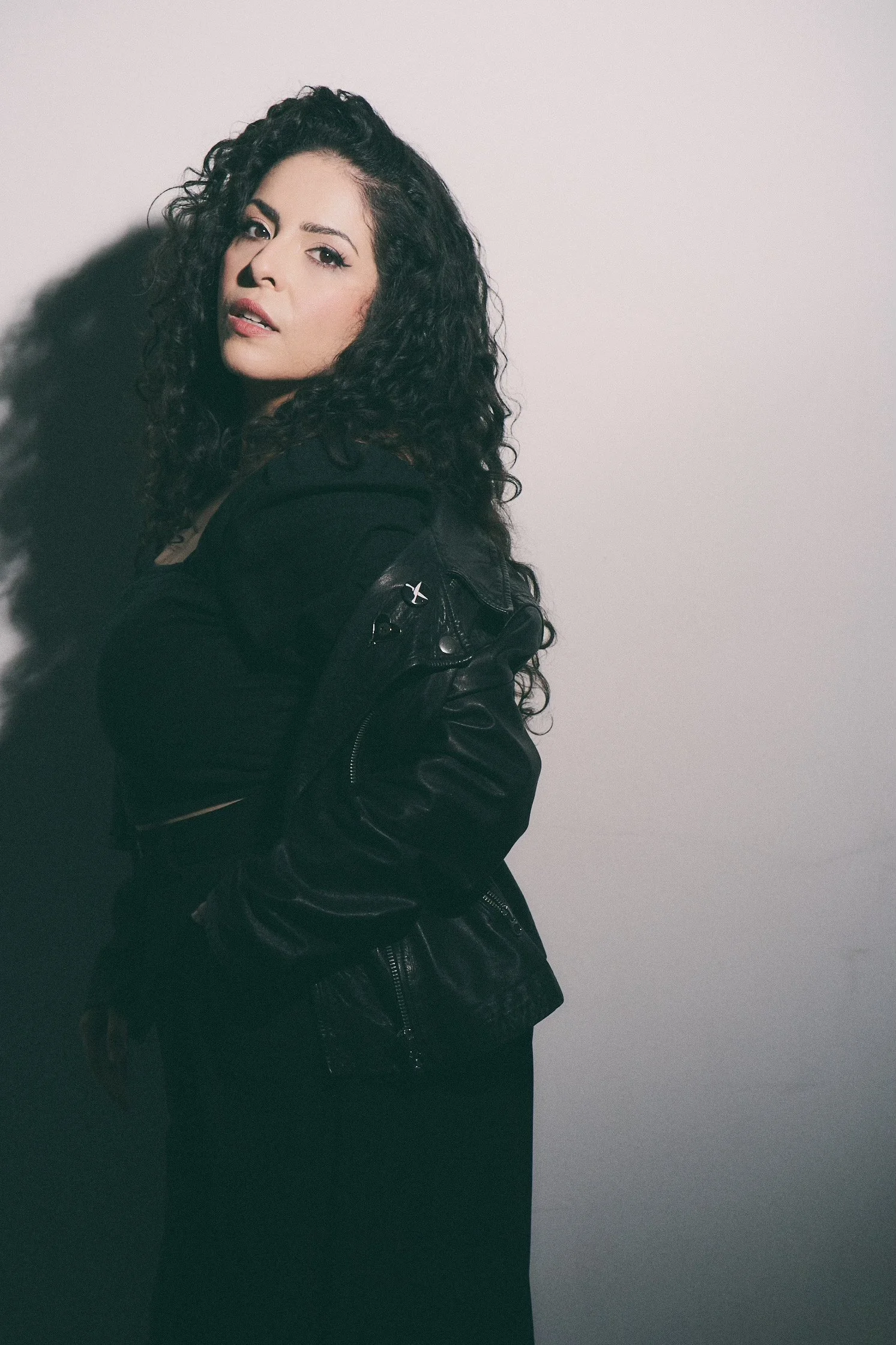 A woman with long, curly dark hair and fair skin posing against a plain gray background. She is wearing a black leather jacket and looking at the camera.