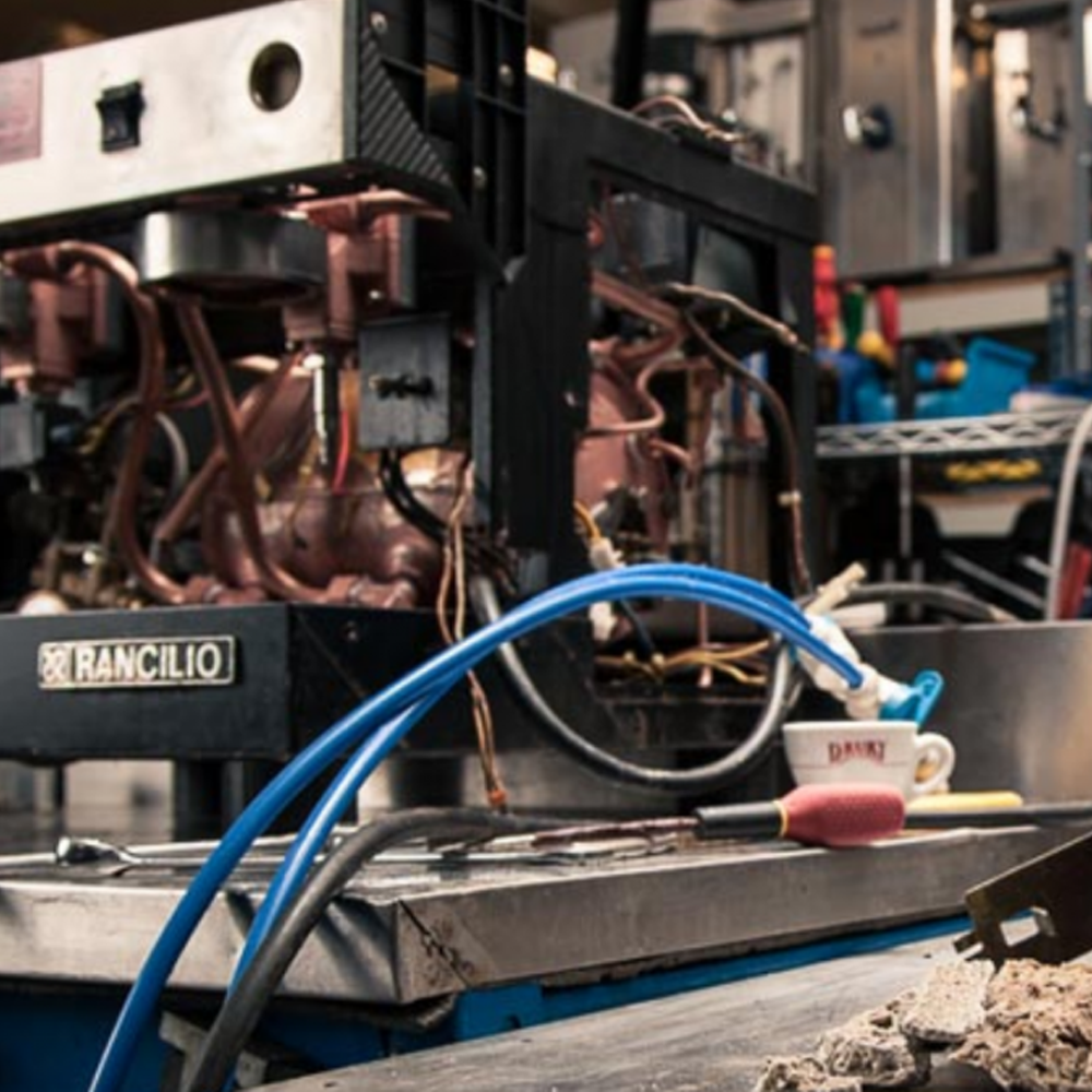 Close-up of mechanical equipment with copper pipes, wires, and tools on a workbench, including a coffee cup with a spoon resting on it.