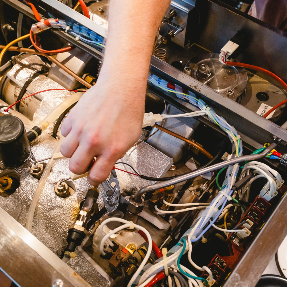 Person working inside a machine, handling wiring and components with various wires, tubes, and metallic parts visible.