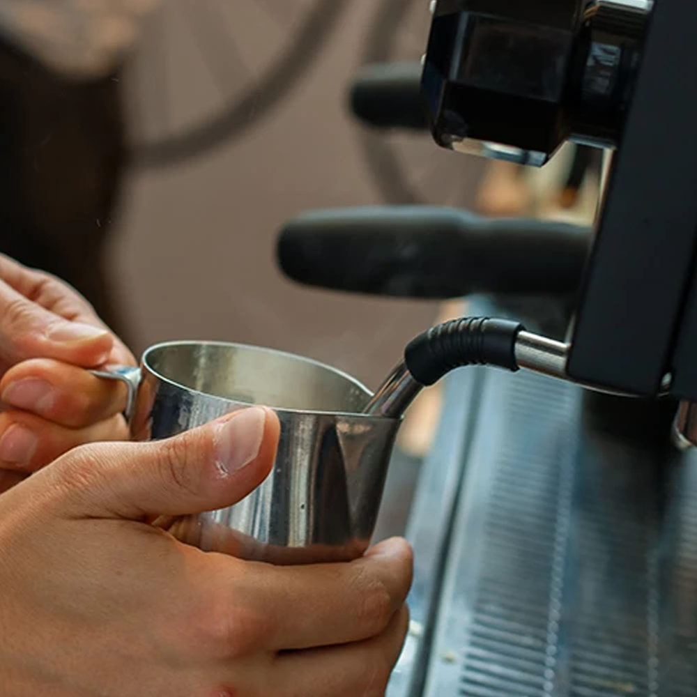Person holding a stainless steel milk frothing pitcher under an espresso machine steam wand for frothing milk.
