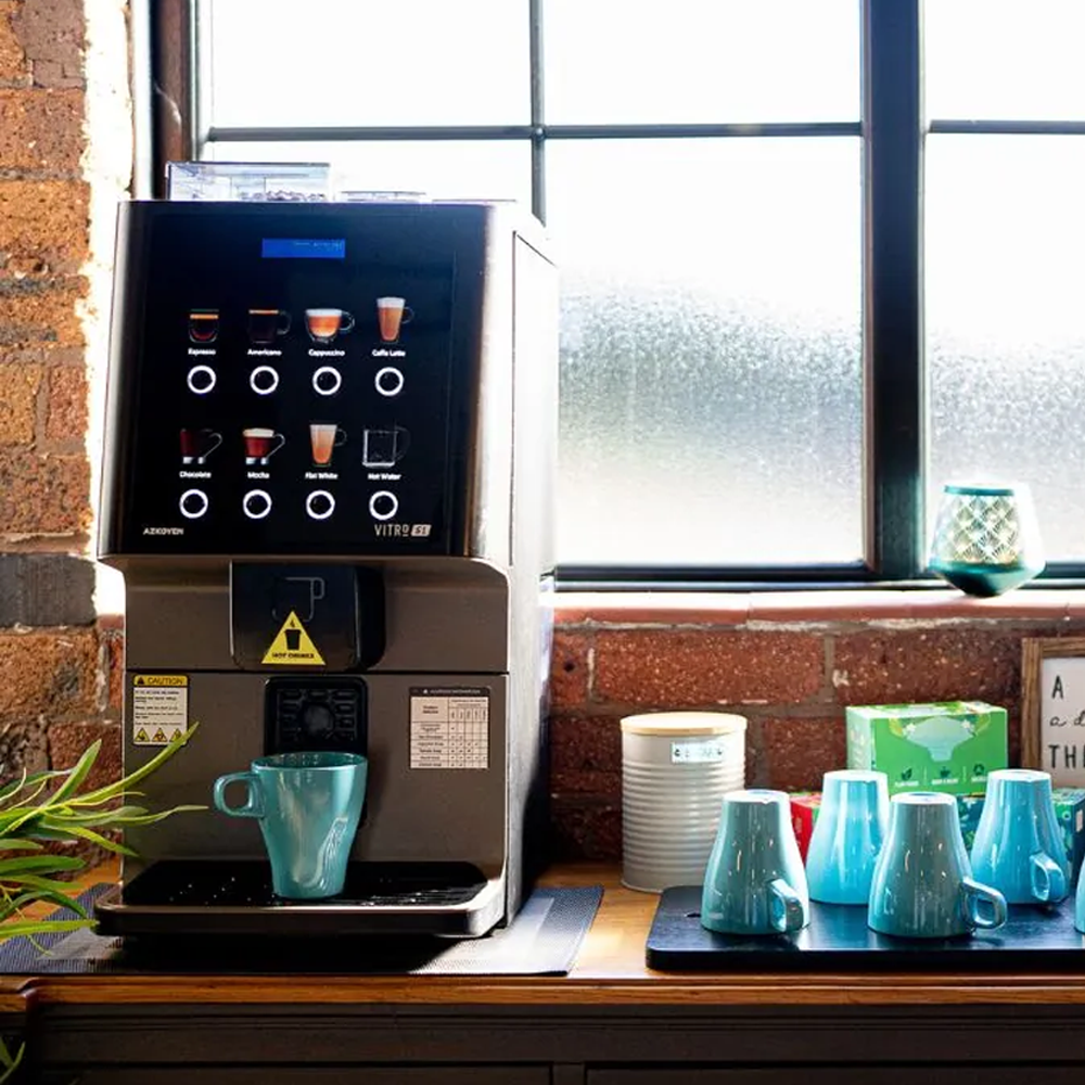 A coffee machine on a wooden counter with a turquoise mug underneath, next to a few more turquoise mugs, and a window in the background.