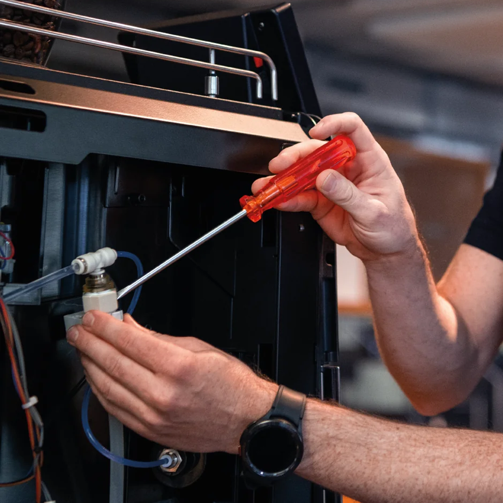 Person repairing or inspecting a black electronic device or machine, using a screwdriver in a workshop or technical setting.