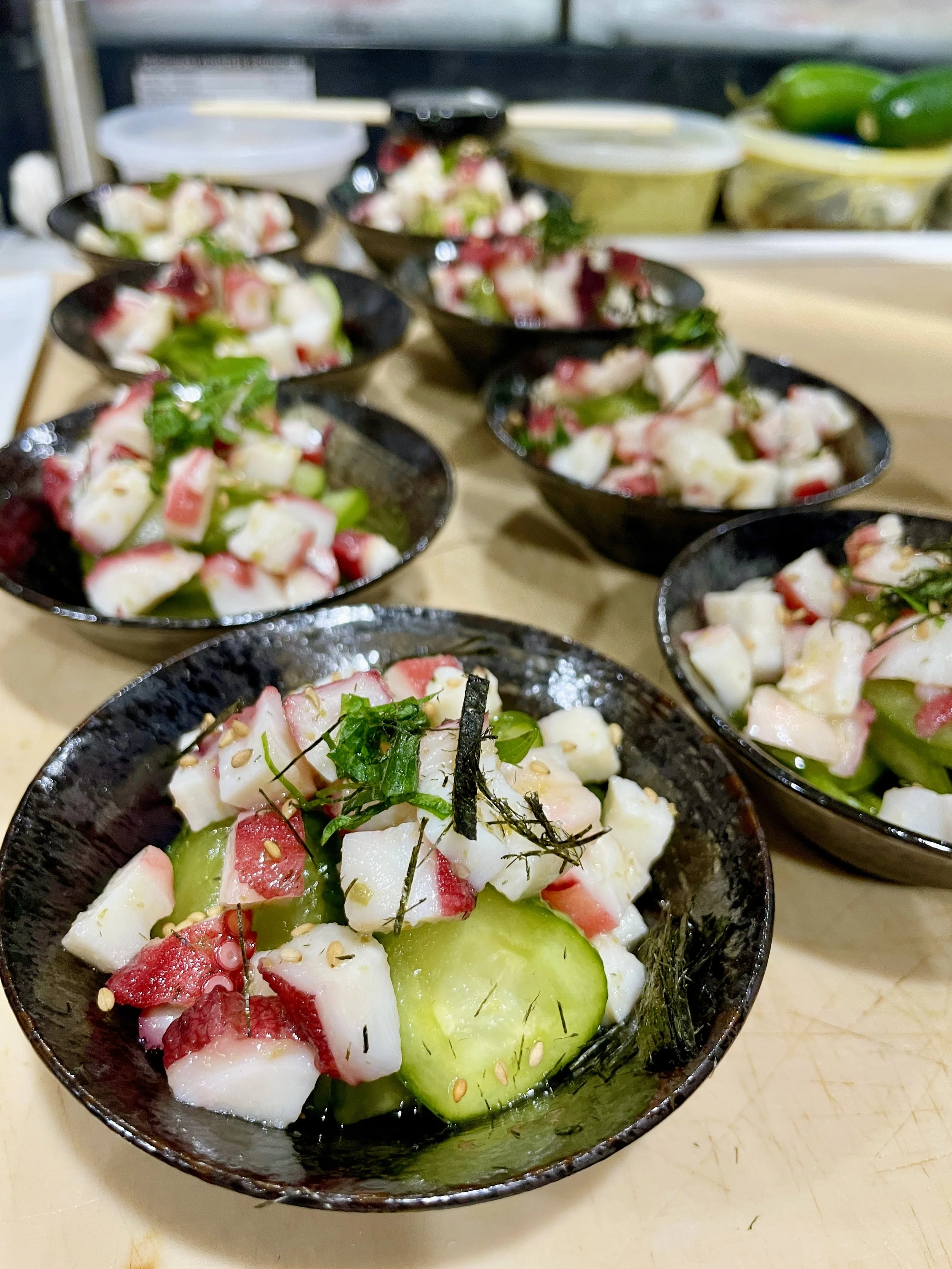 Small black bowls filled with cubed pieces of octopus, sliced cucumber, red radish, and garnished with herbs and sesame seeds. The bowls are arranged on a wooden surface.