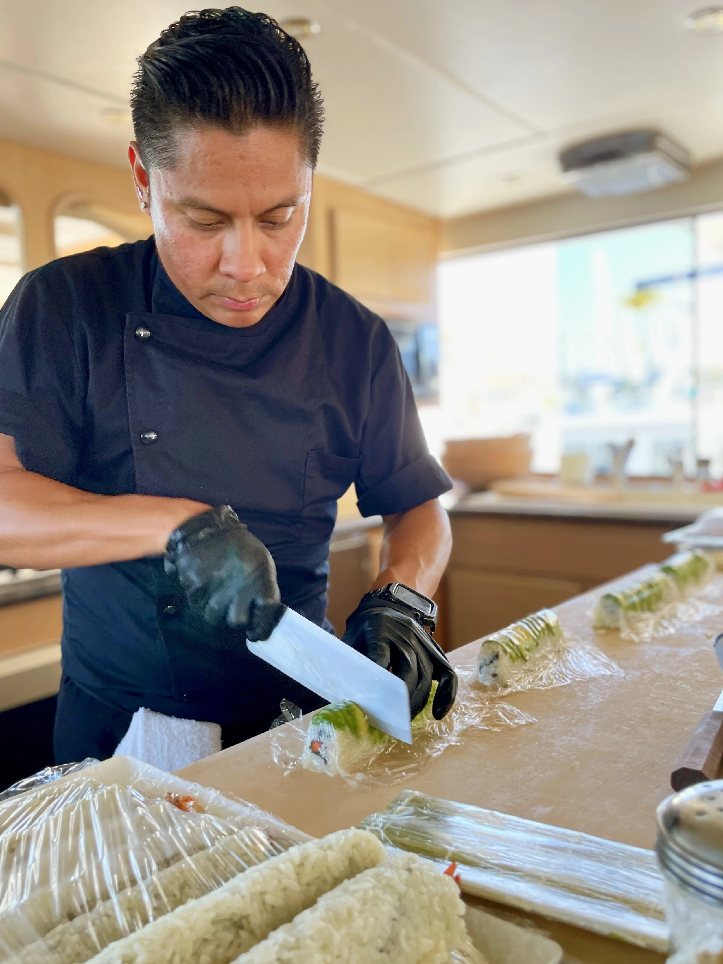 A chef in a black uniform and gloves slicing sushi rolls on a wooden table covered with plastic wrap, with fresh sushi ingredients and rice in the foreground.