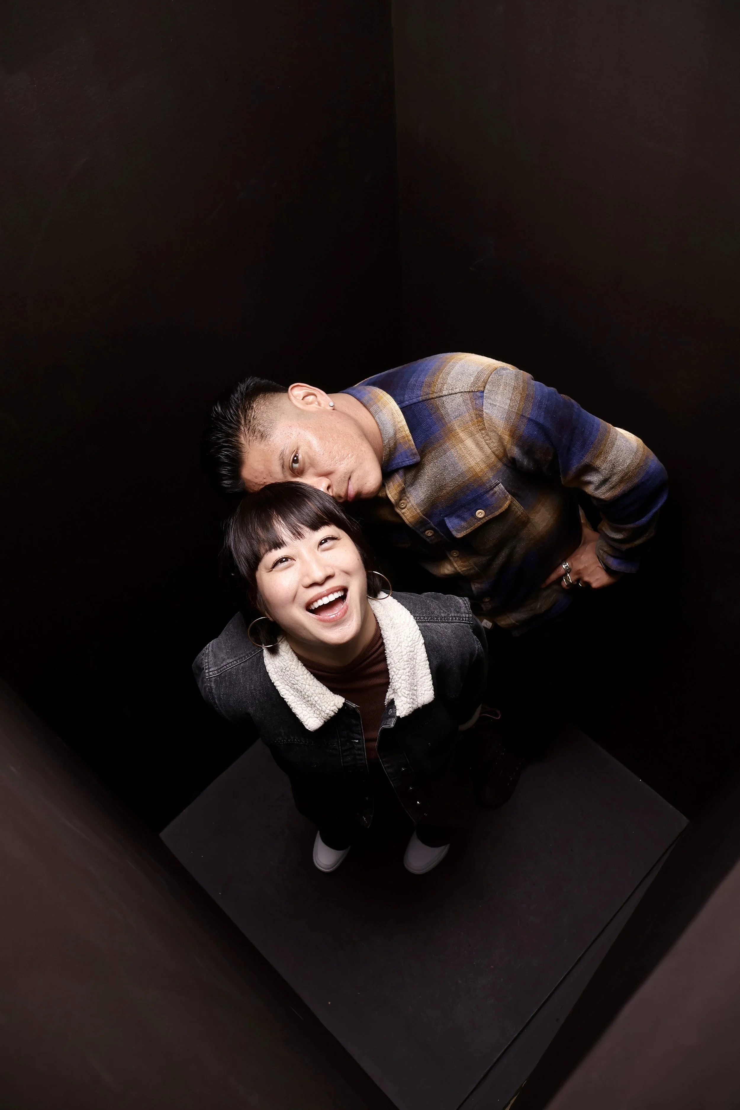 Two people, a man and a woman, posing closely together inside a dark, reflective elevator. The woman is smiling widely at the camera, wearing a dark jacket with a sherpa collar and hoop earrings. The man is looking at the camera with a serious expression, dressed in a plaid shirt layered over a darker shirt.