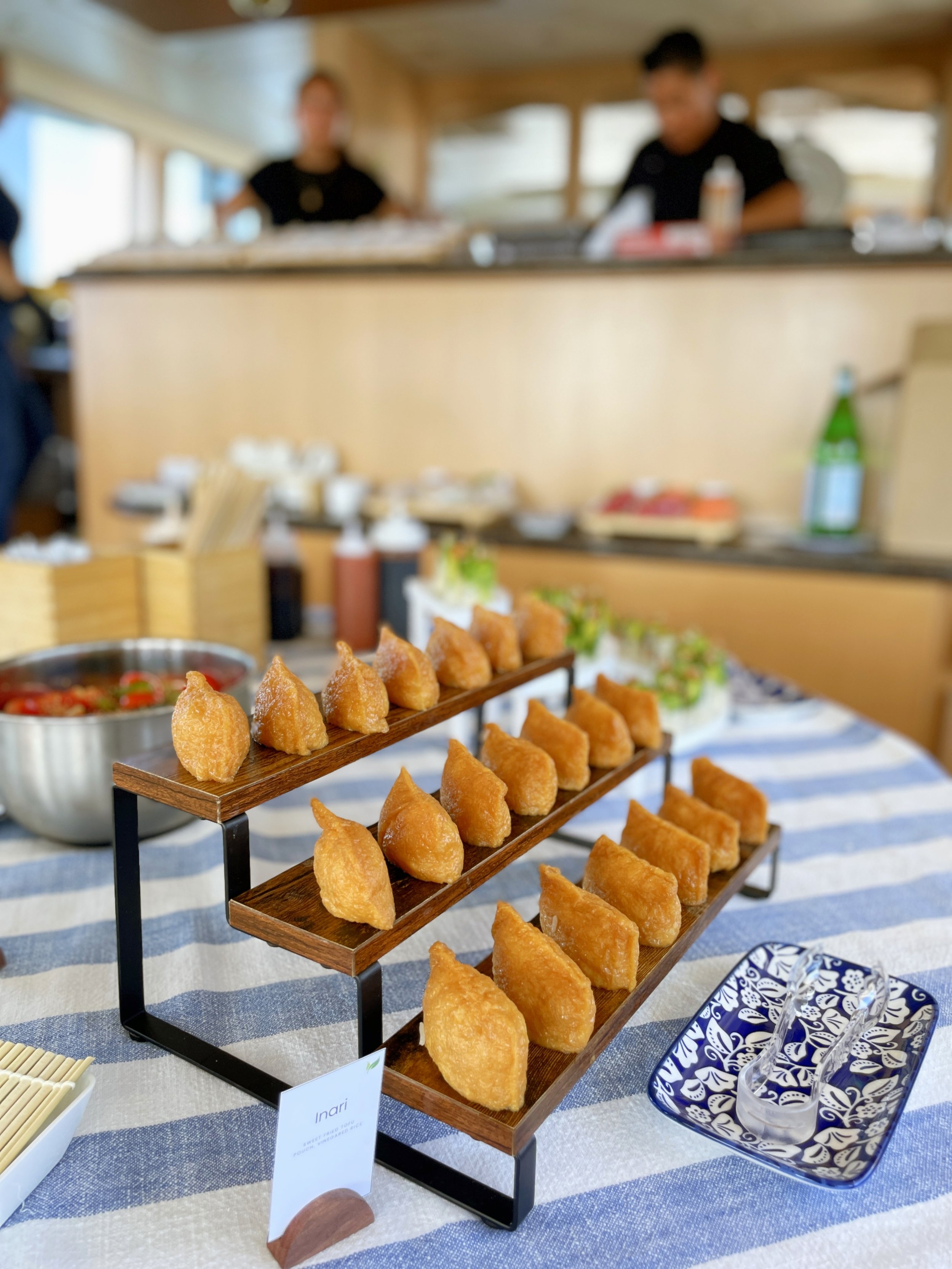 Close-up of a tiered wooden and metal stand filled with fried food, with a small bowl of spicy sauce on the side, set on a blue-and-white striped tablecloth at a buffet or food presentation.