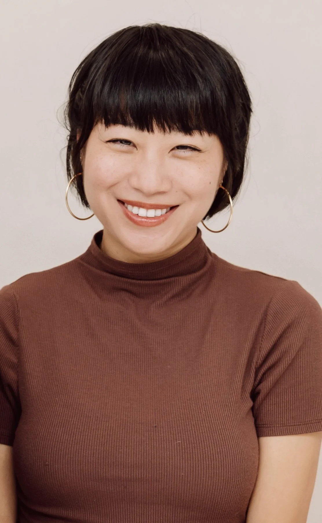 A woman with short dark hair, wearing large hoop earrings and a high-neck brown shirt, smiling at the camera against a plain light background.
