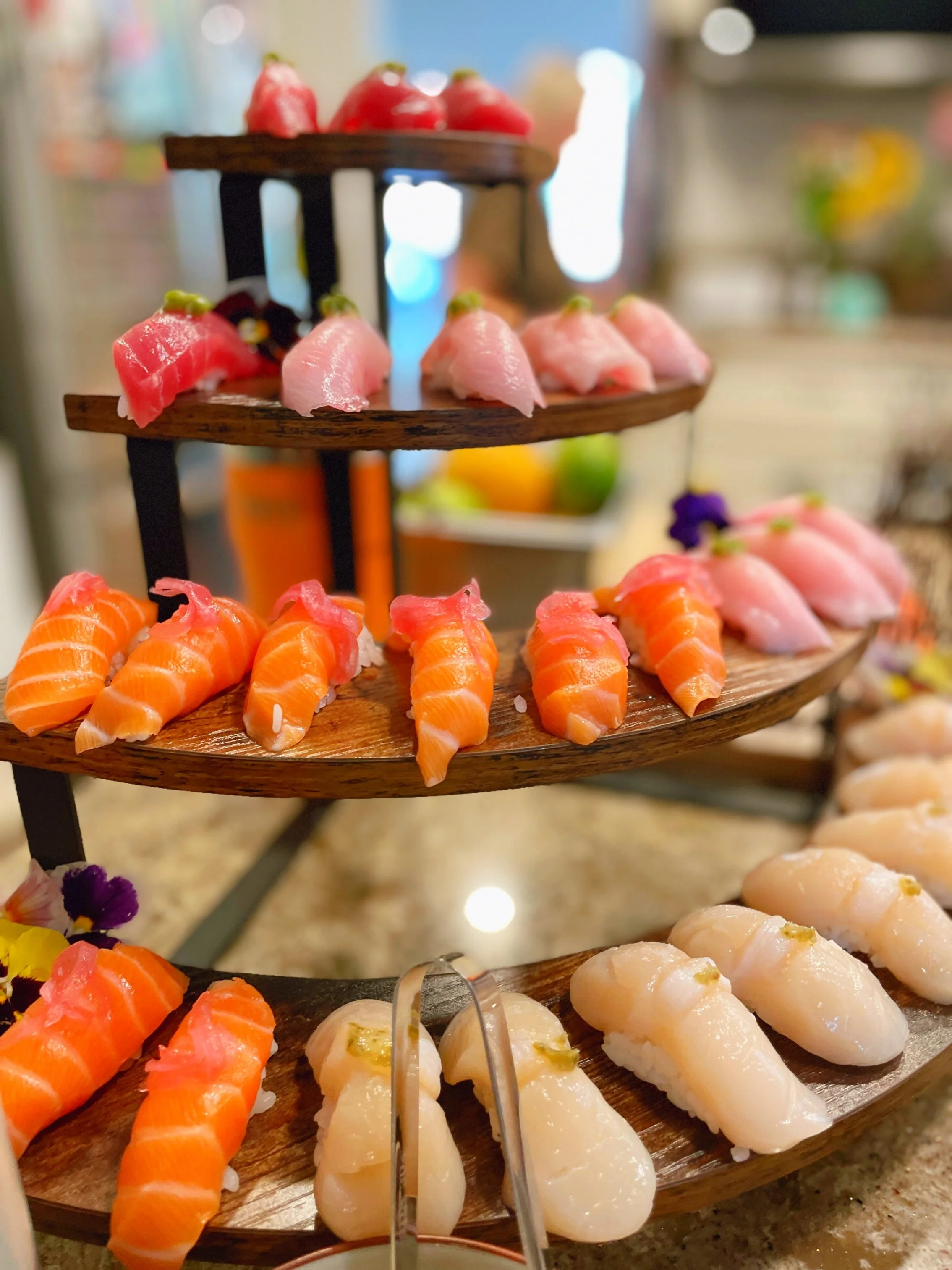 A multi-tiered wooden sushi platter with various types of nigiri, including salmon, tuna, and white fish, arranged on different levels, with some garnishes and flowers around it.