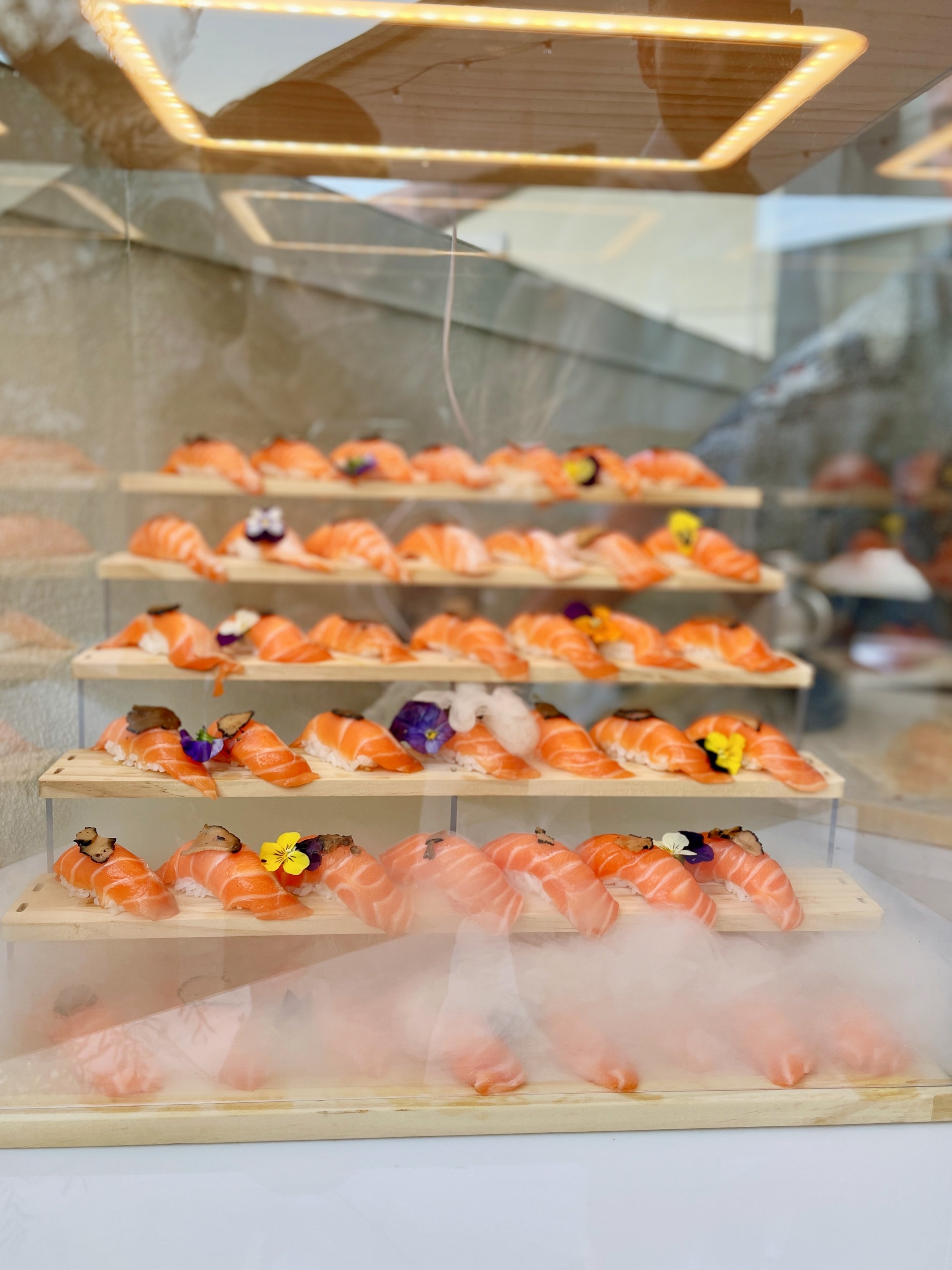 Multiple trays of fresh salmon nigiri garnished with edible flowers and herbs, displayed on wooden platforms inside a glass display case at a private event.