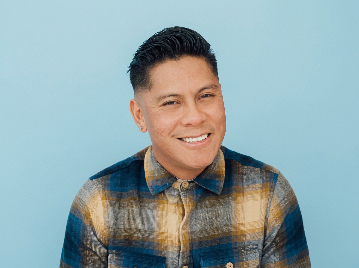 A young man with dark, slicked-back hair smiling in front of a light blue background, wearing a plaid shirt.