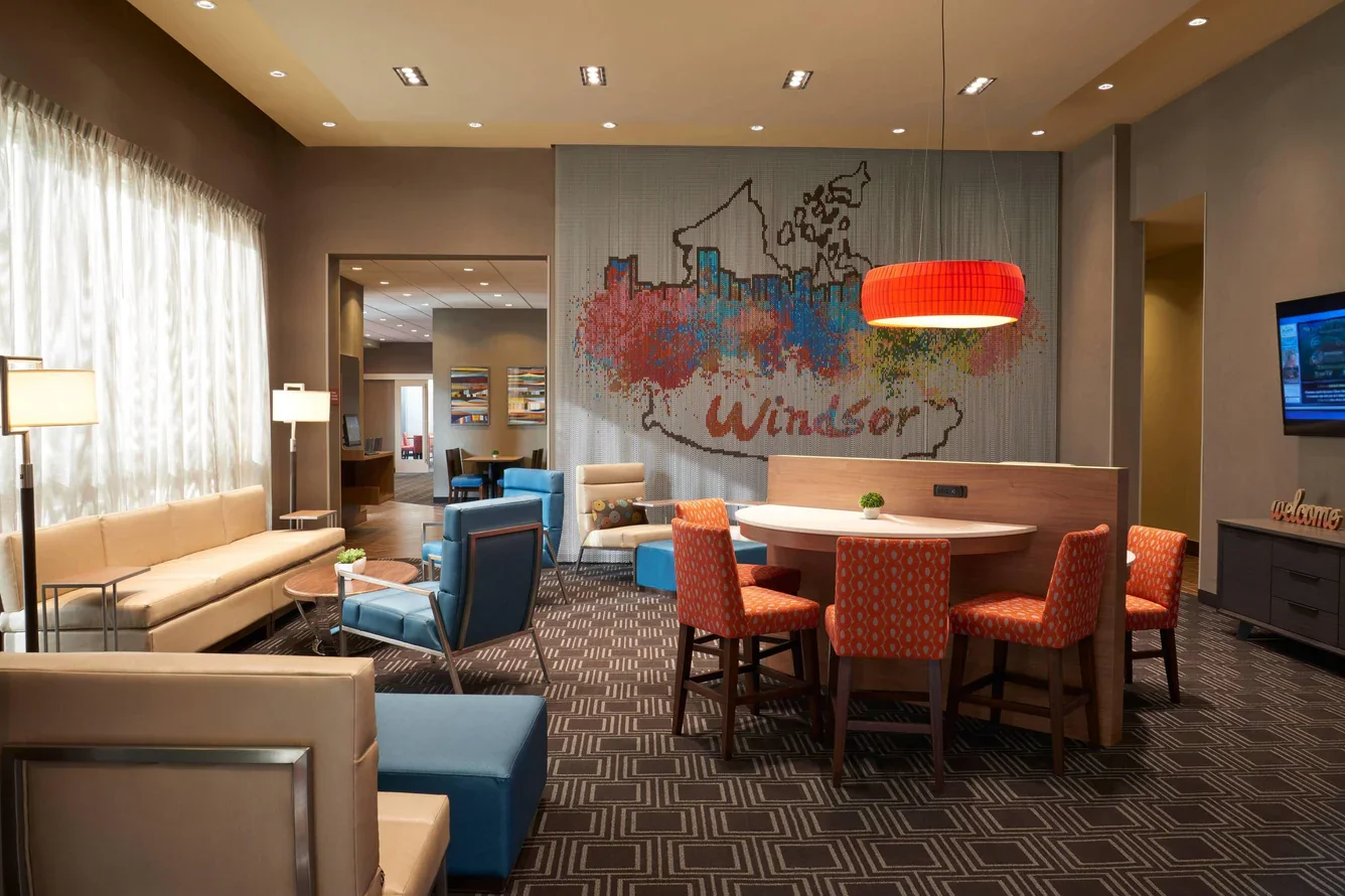 Hotel lobby with seating area including sofas, armchairs, and a group of four orange patterned chairs around a white round table. Large window with curtains, wall with colorful artwork, and a red pendant light.