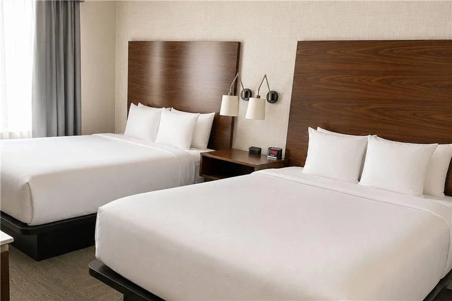 Two queen-sized beds with white linens and pillows in a hotel room, wooden headboards, nightstand with lamps, clock, and phone, beige walls, and a window with gray curtains.