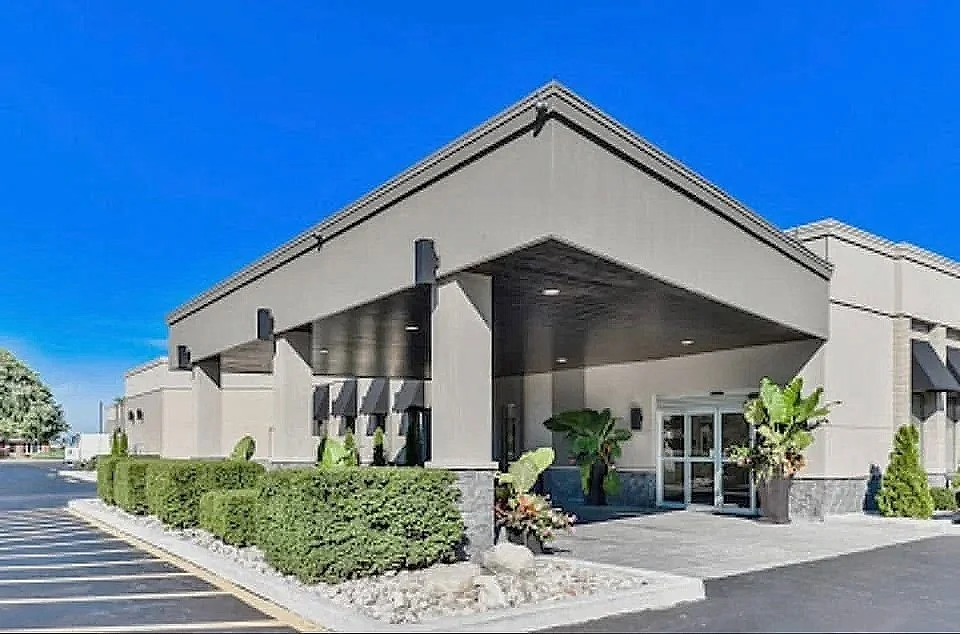 Modern commercial building with a covered entrance, well-maintained bushes, large potted plants, and a parking lot under a clear blue sky.