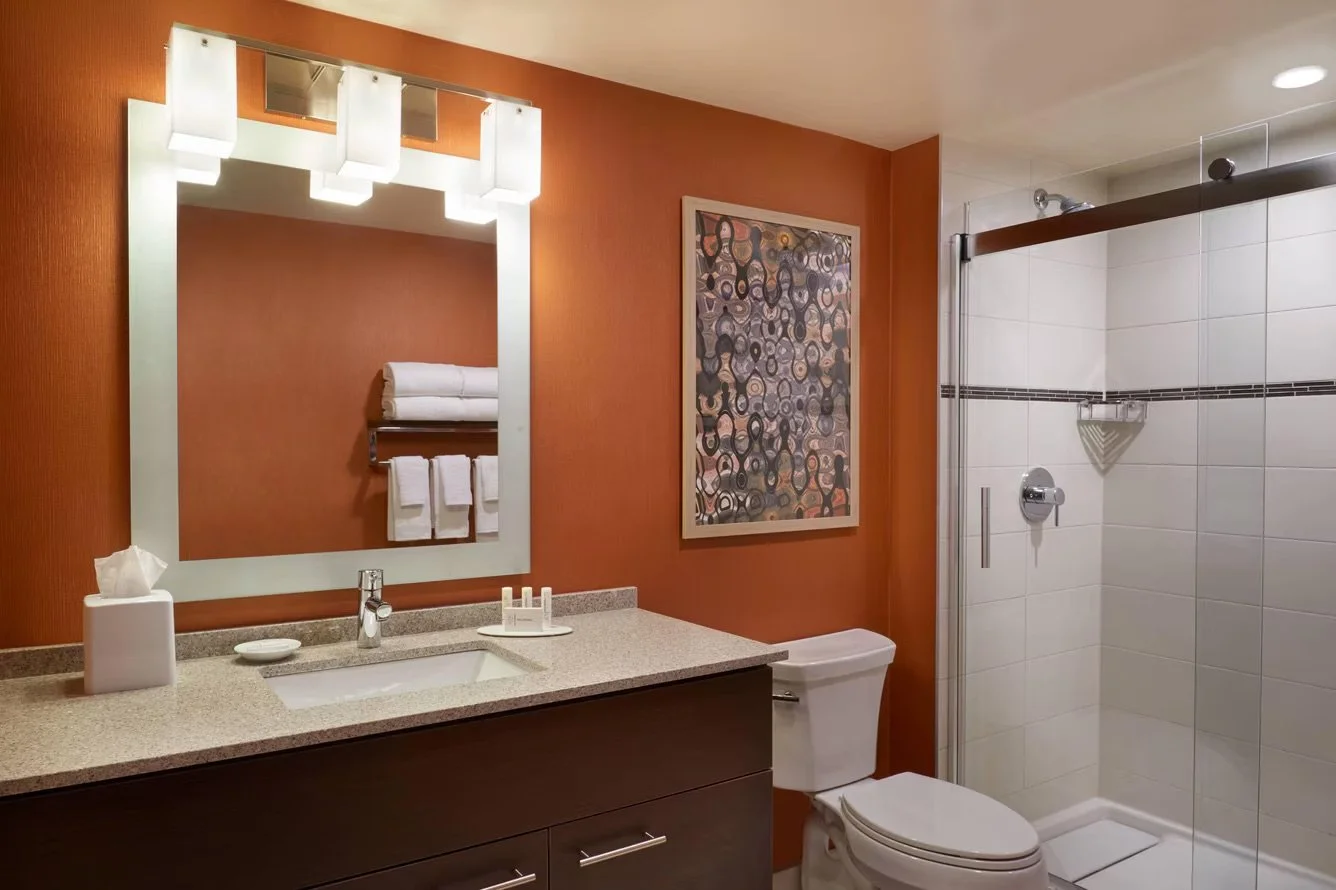 Bathroom with a large mirror, sink with toiletries, toilet, glass shower enclosure, and a colorful abstract painting on orange wall.
