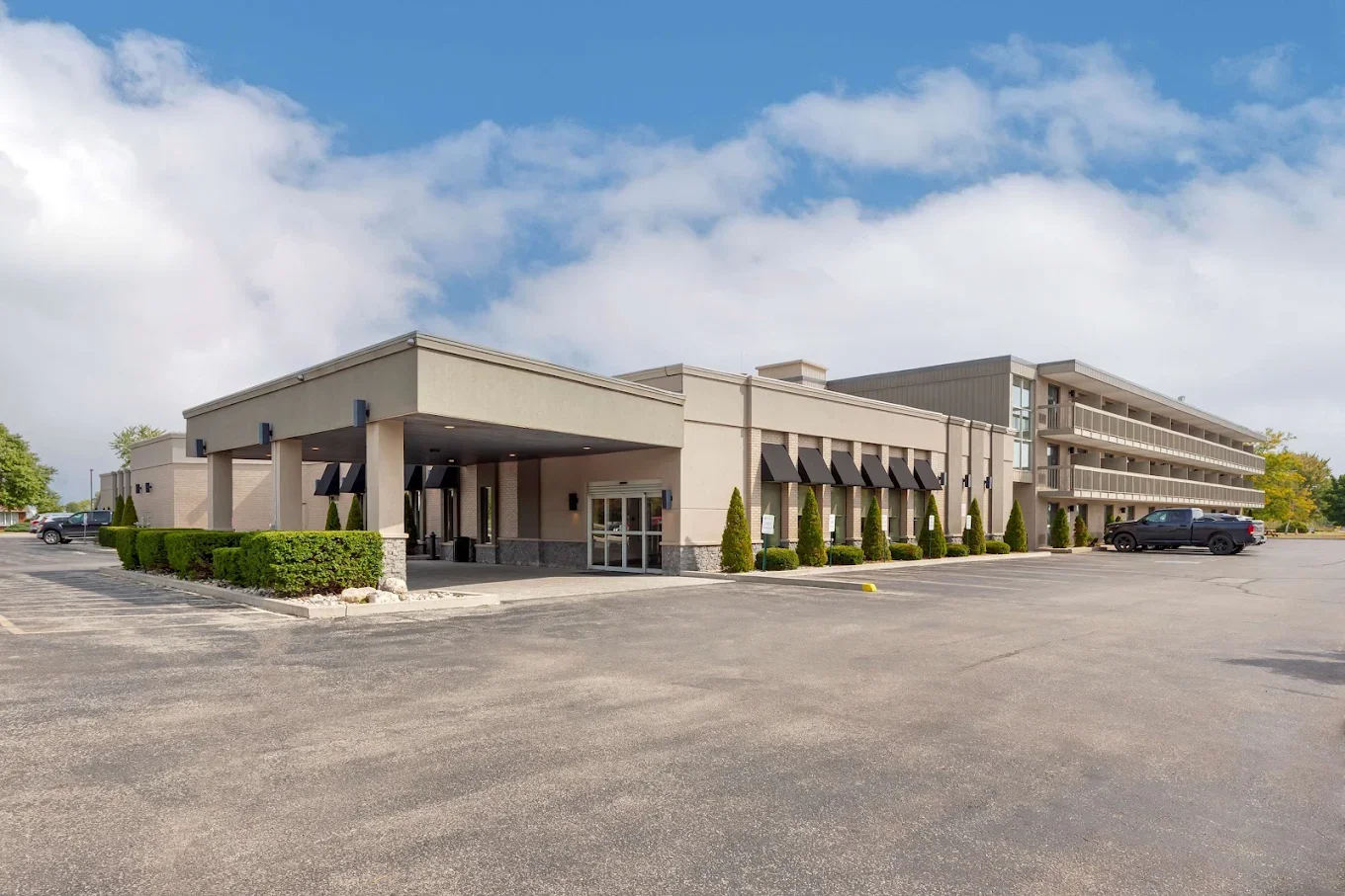 A three-story motel with a beige exterior and black awnings, surrounded by a parking lot with a few parked cars and trimmed bushes, under a partly cloudy sky.