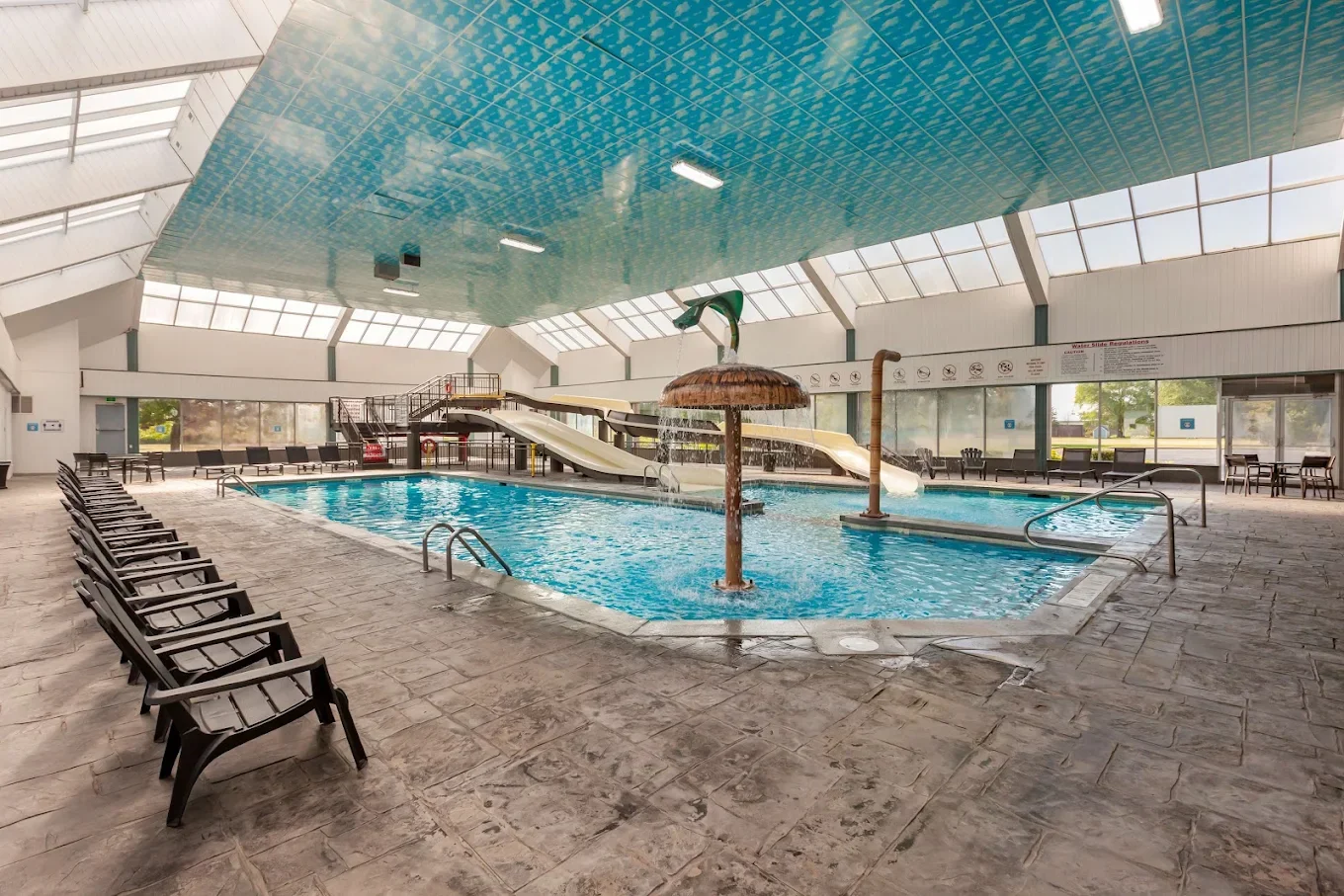 Indoor swimming pool with multiple water slides, a mushroom-shaped water fountain, and lounge chairs around the pool area, under a glass ceiling.