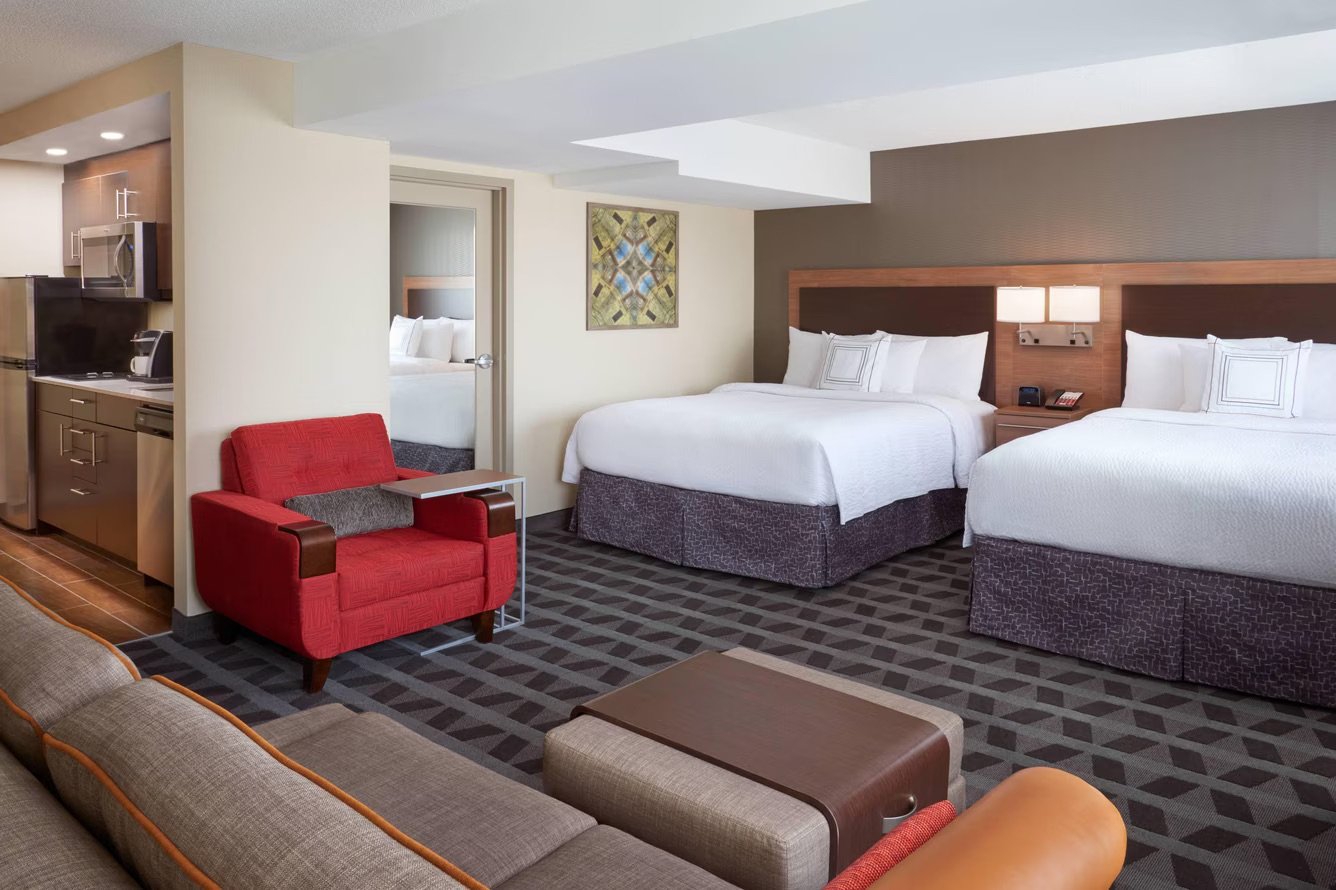 Hotel room with two queen beds, a sitting area with a red armchair and a gray sofa, and a kitchenette in the corner.